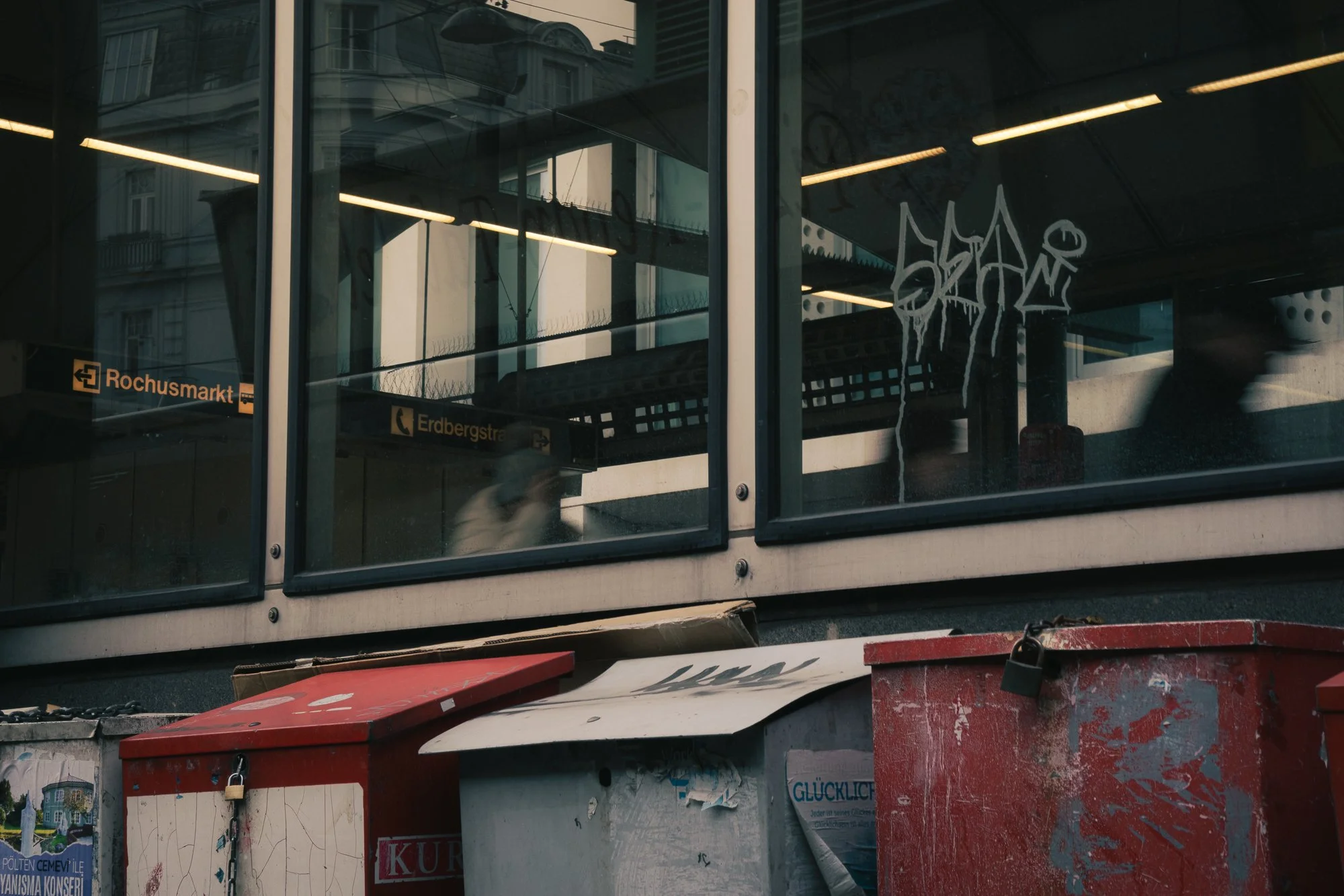 Reflections of buildings and street signs visible through glass panels of a building's exterior. The street signs read 'Rochusmarkt' and 'Erdbergstraße'. Graffiti is present on the glass. In the foreground, there are red and white trash and recycling