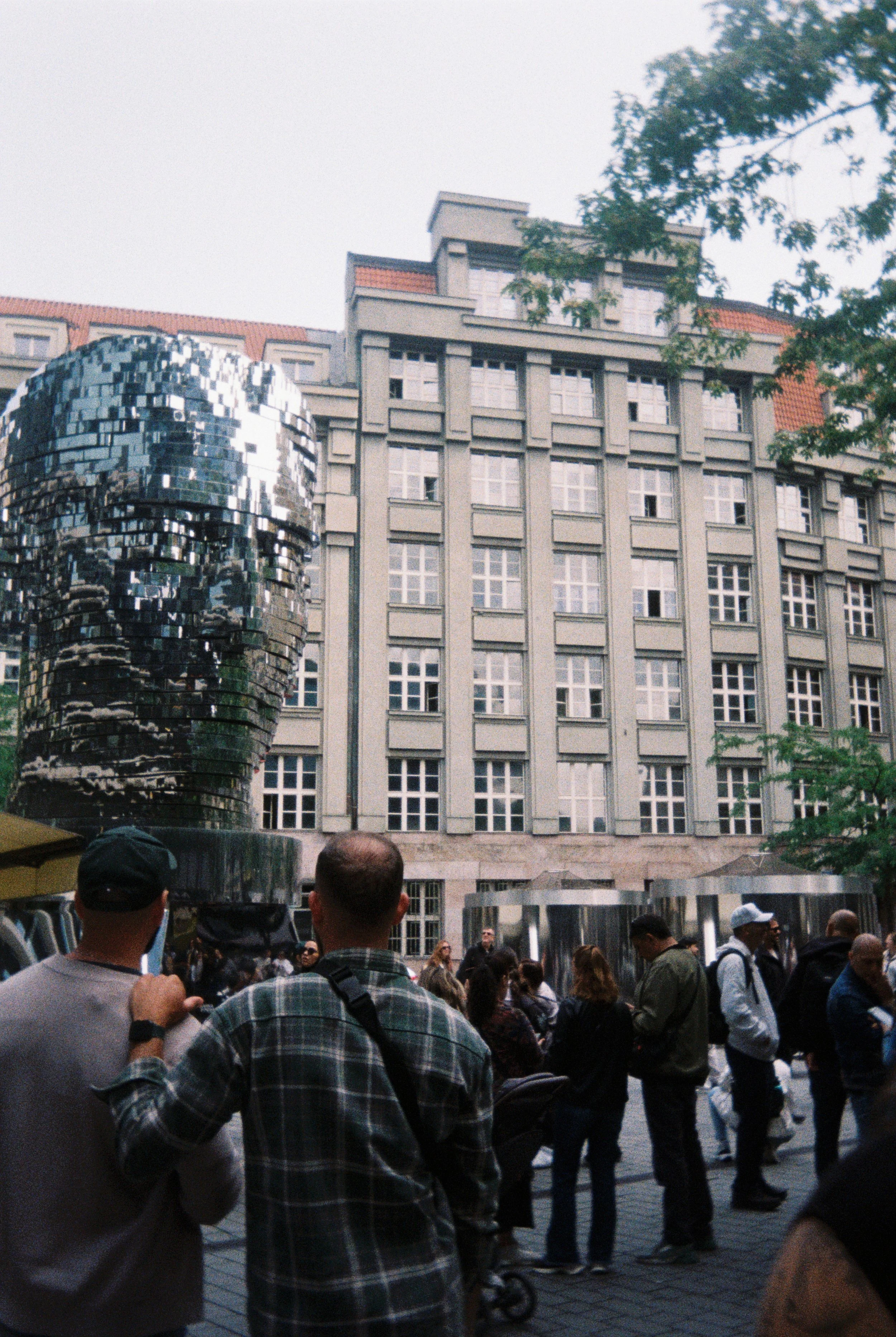 A busy outdoor scene with a crowd of people in front of a large concrete building with many windows. There is a shiny, reflective sculpture on the left side of the image and some trees on the right and upper part of the scene.