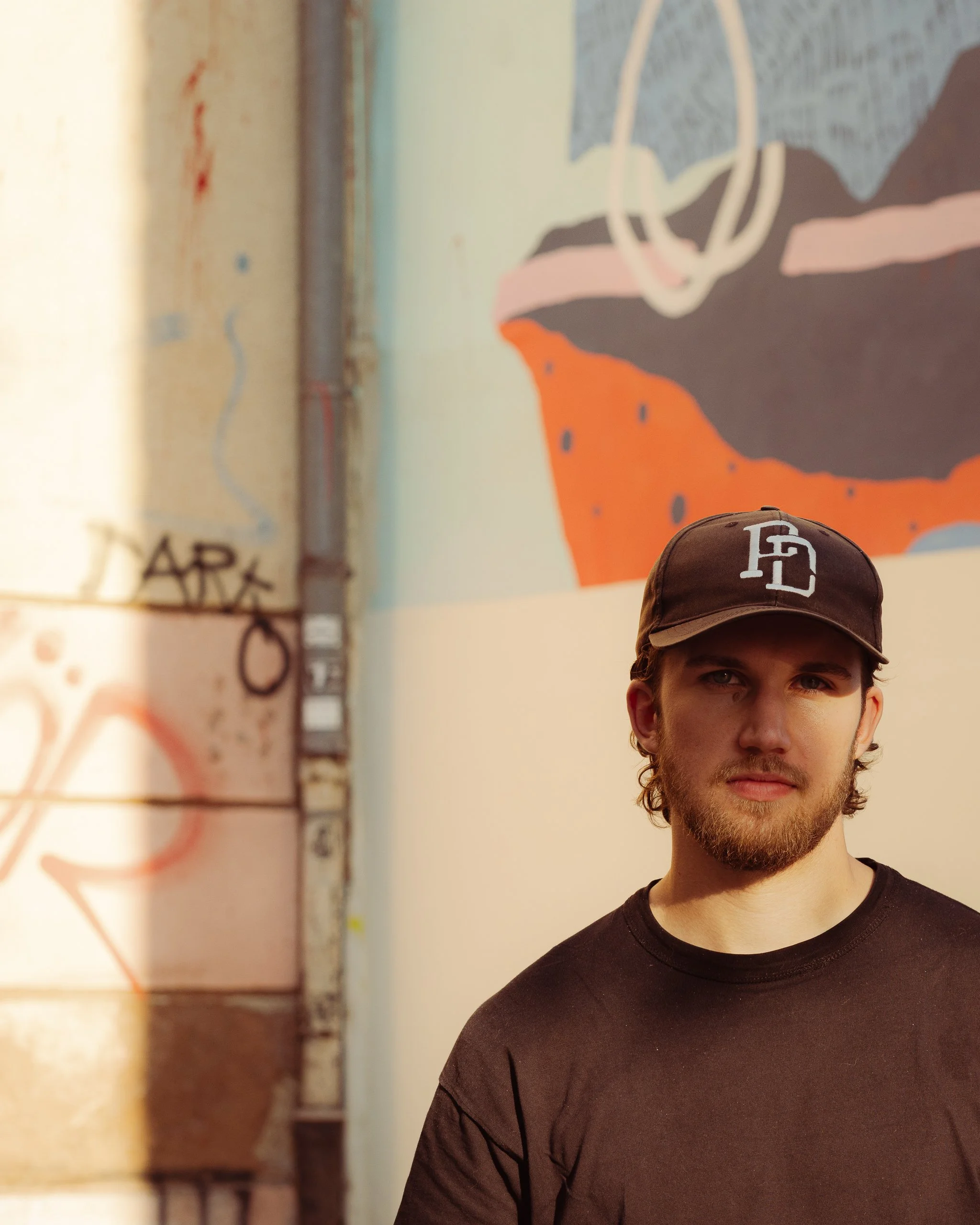 A young man with a beard and wearing a dark baseball cap with a logo stands indoors in front of a colorful abstract mural with mountain shapes, large loops, and splashes of color.