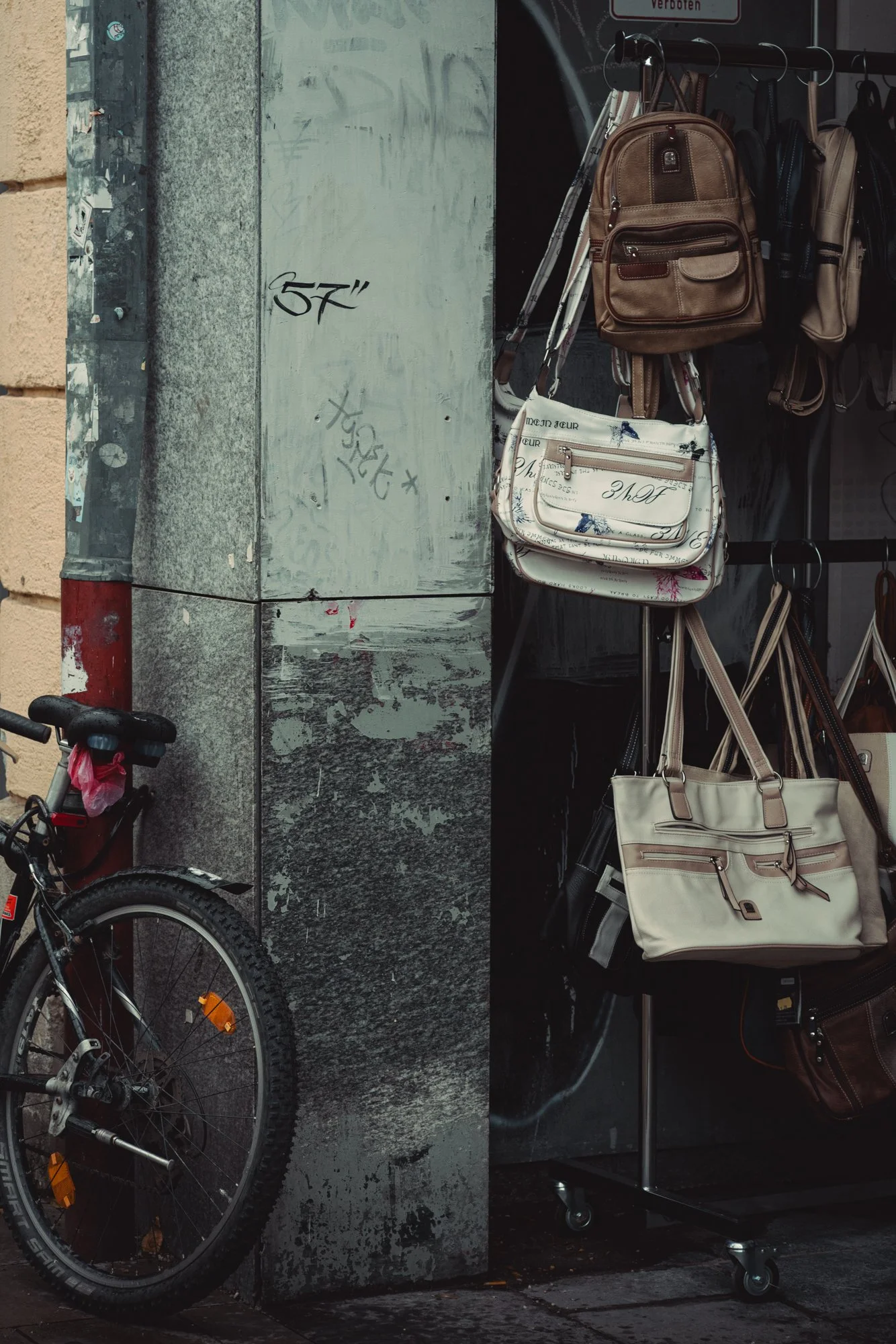 A display of handbags and backpacks hanging on a rack outside a store, with a bicycle parked nearby and a worn utility box with graffiti on it.