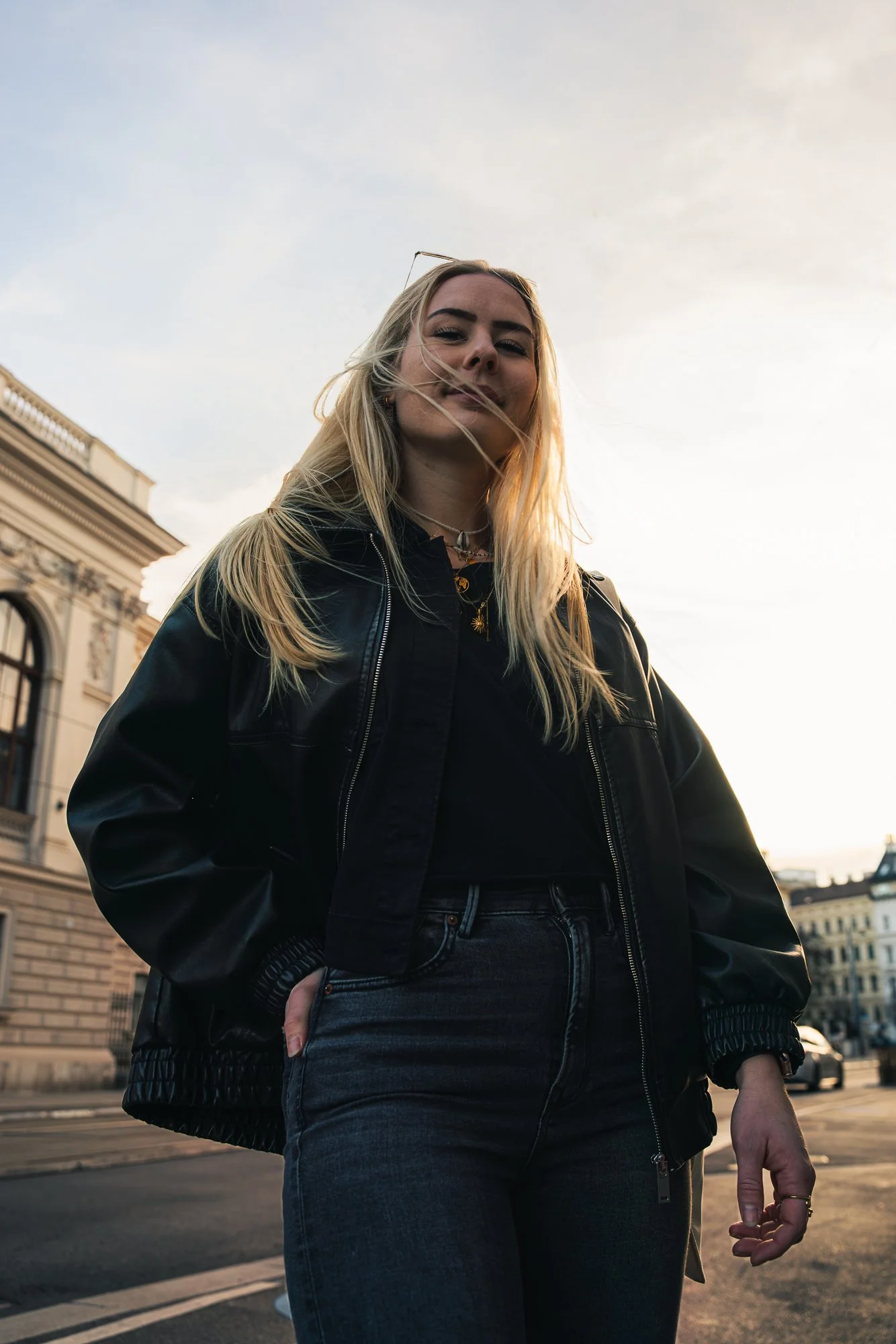 A young woman with long blonde hair wearing a black leather jacket and dark jeans standing outdoors on a city street during sunset, with historic buildings in the background.