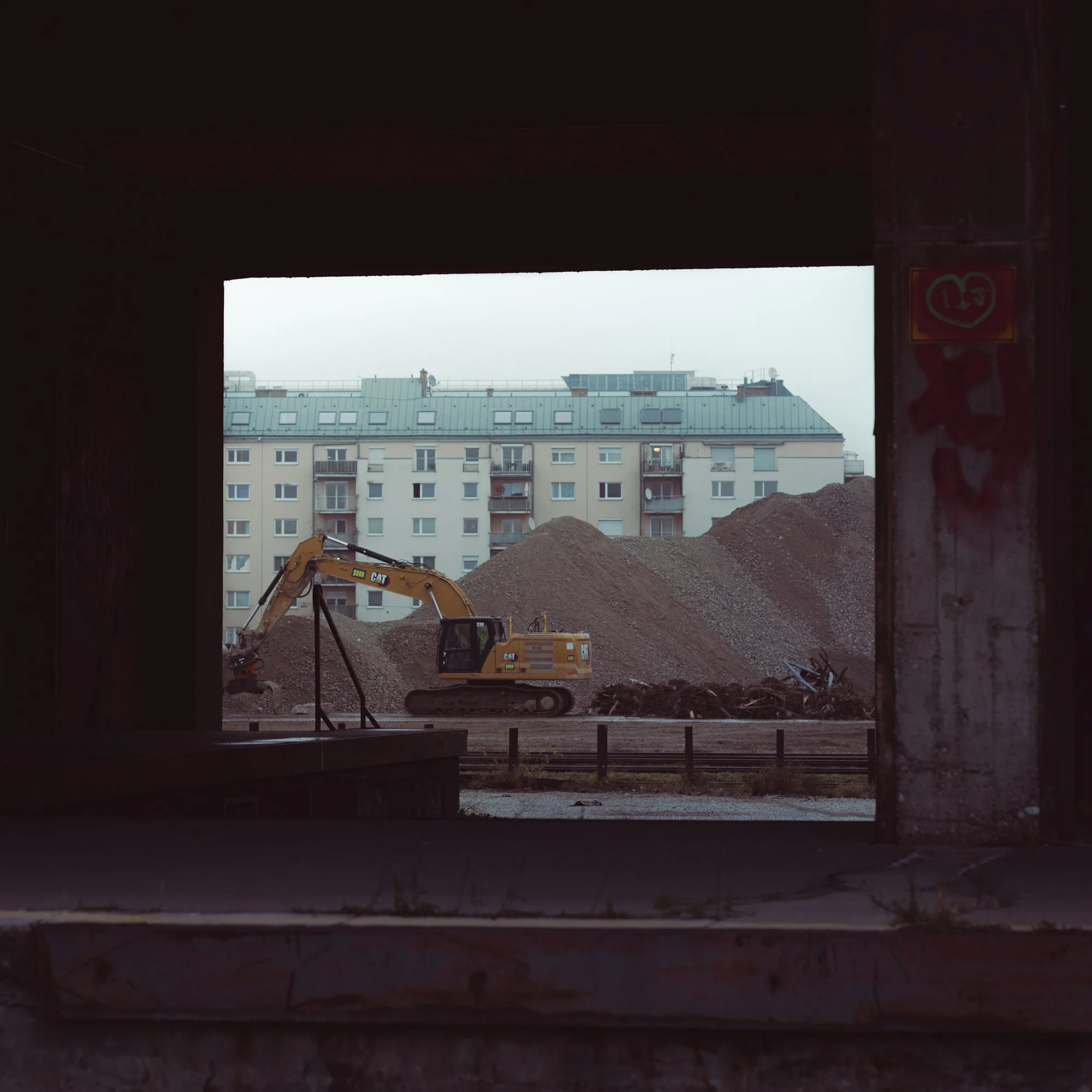 Construction site with excavator and dirt mound viewed through a dark opening, with residential buildings in the background.