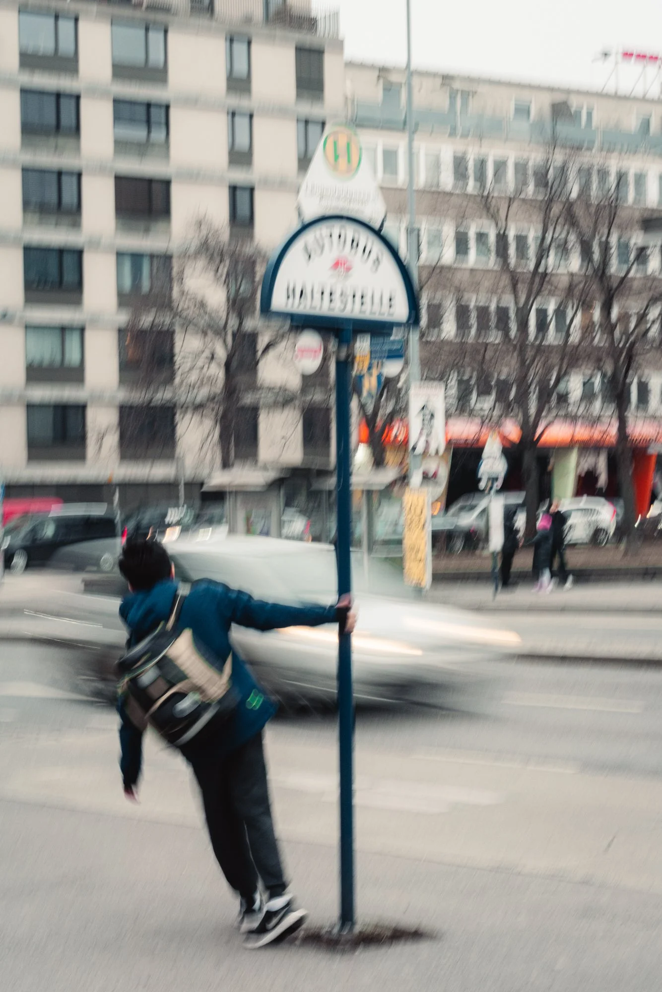 A person in a blue jacket and black pants is falling or losing balance while running or walking quickly on the sidewalk near a street in an urban area. In the background, there are multiple cars, a building with multiple floors, and pedestrians.