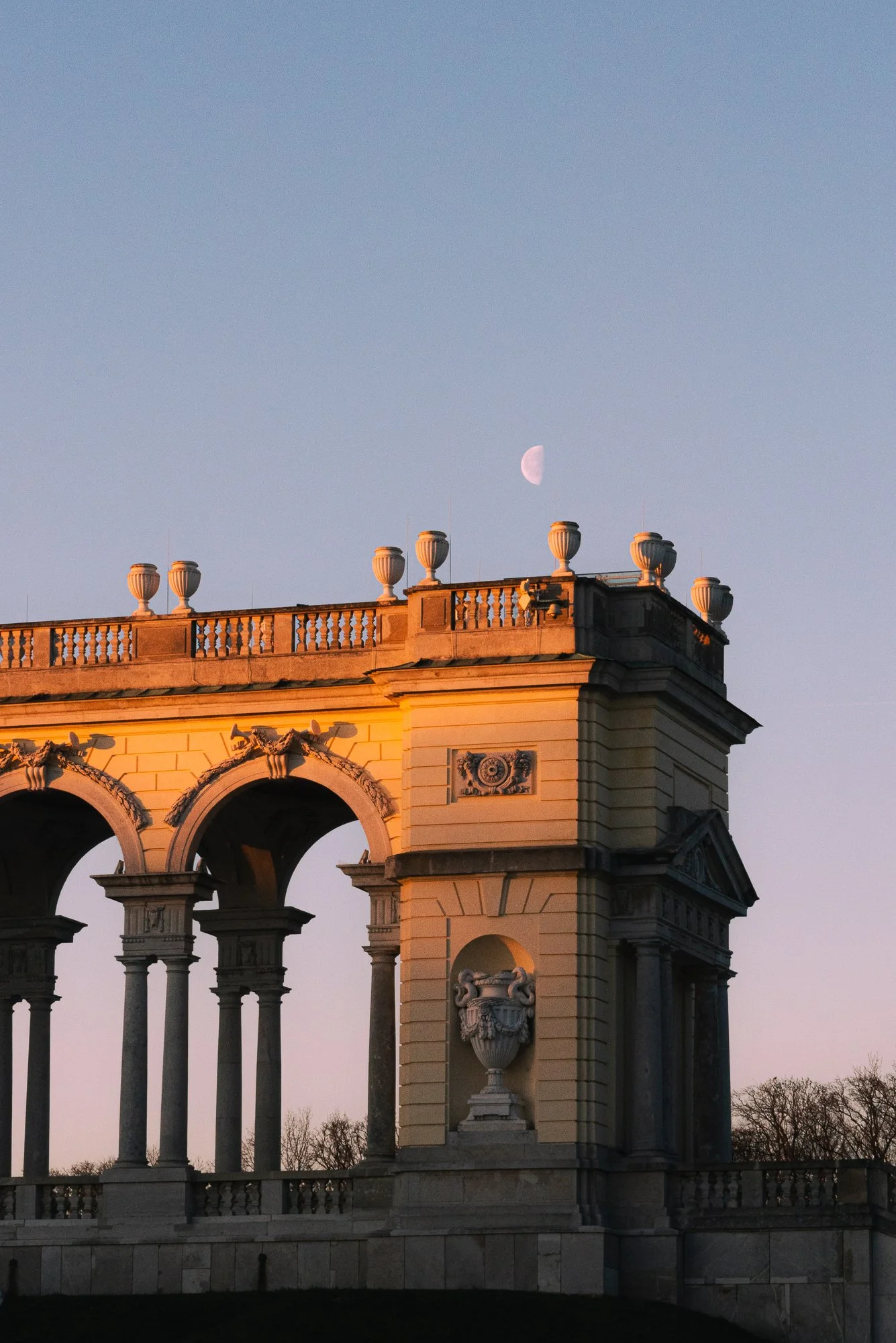 A historic building with tall arches, decorative urns on the rooftop, and sculptures on its facade, illuminated by warm sunlight with the moon visible in the sky.