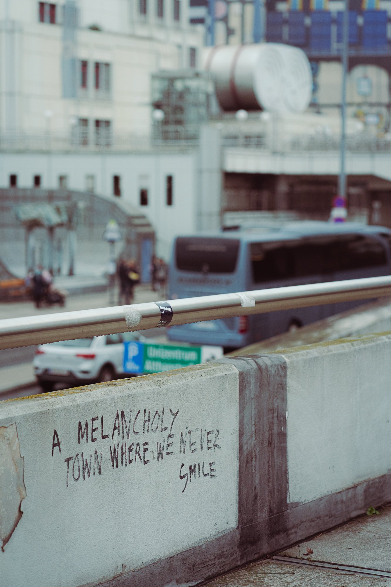 A view from a balcony showing cars, pedestrians, buildings, and a street sign that says 'University.' There is a white wall with handwritten text that says, 'A Melancholy town where we never smile' in the foreground.