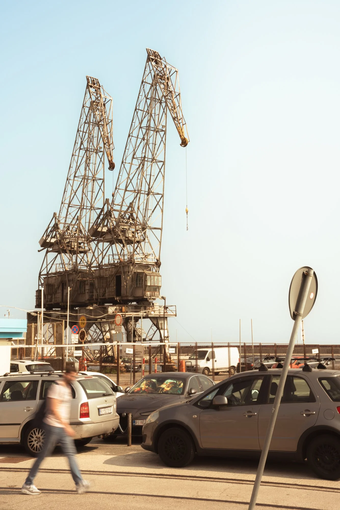 A street scene with a parking lot filled with cars, a person walking by, and a large industrial crane in the background under a clear sky.
