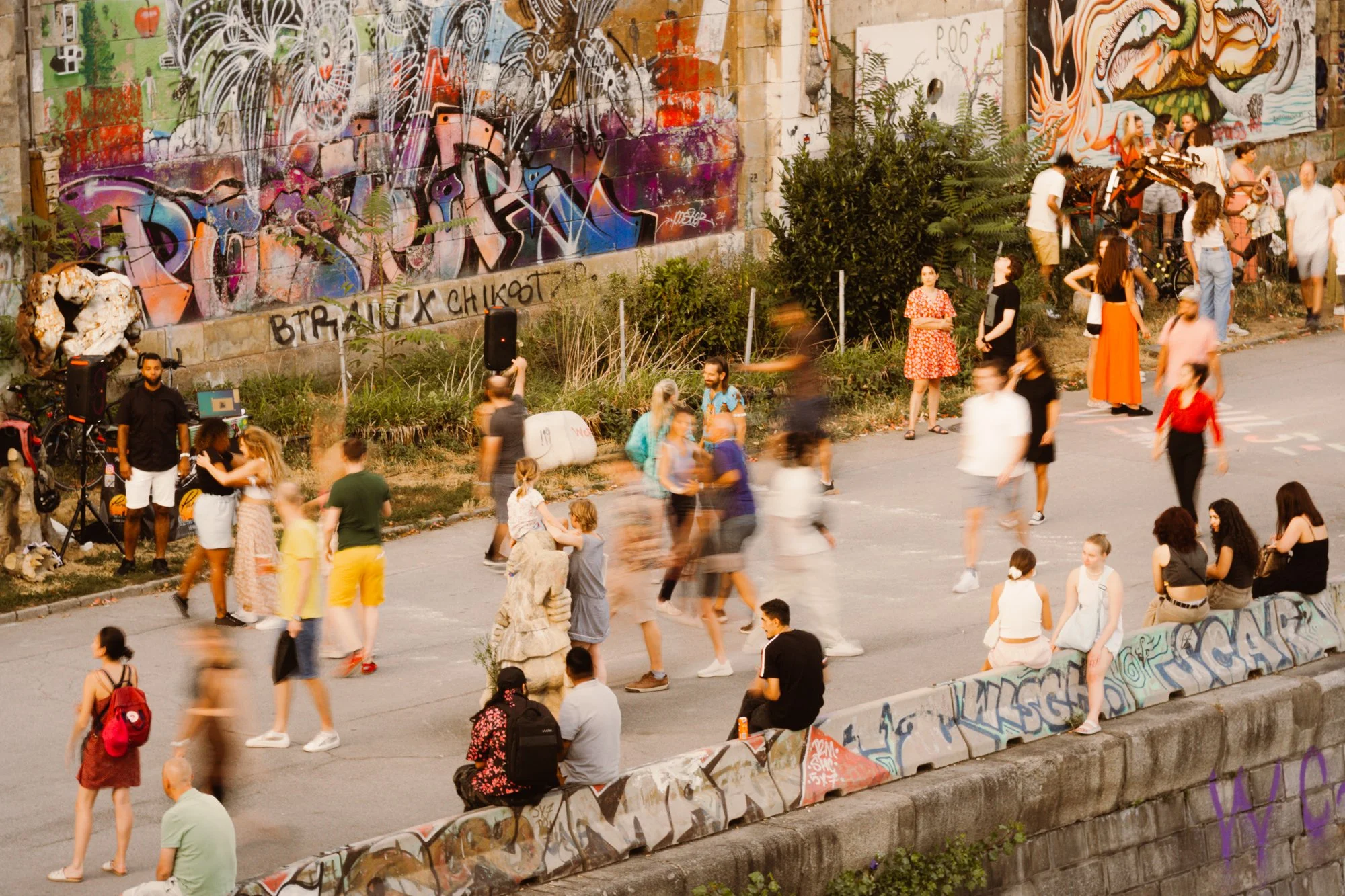 People gather around a graffiti-covered wall on a sidewalk in an urban park.