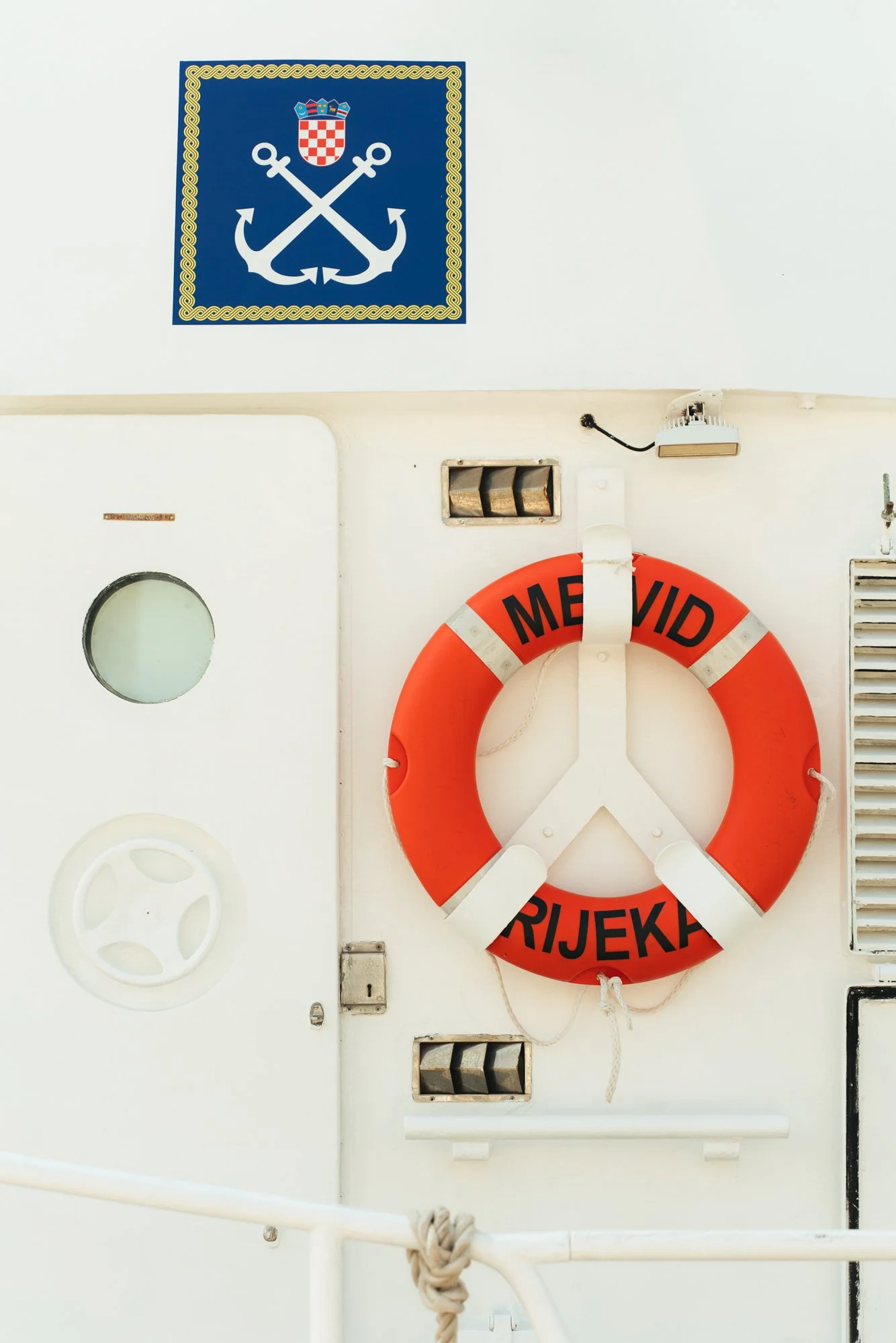 A white wall with a maritime emblem of two crossed anchors, a Croatian coat of arms and a decorative border at the top left corner. A bright orange life ring with black text hangs on the wall, partially covering some vents and a small window.