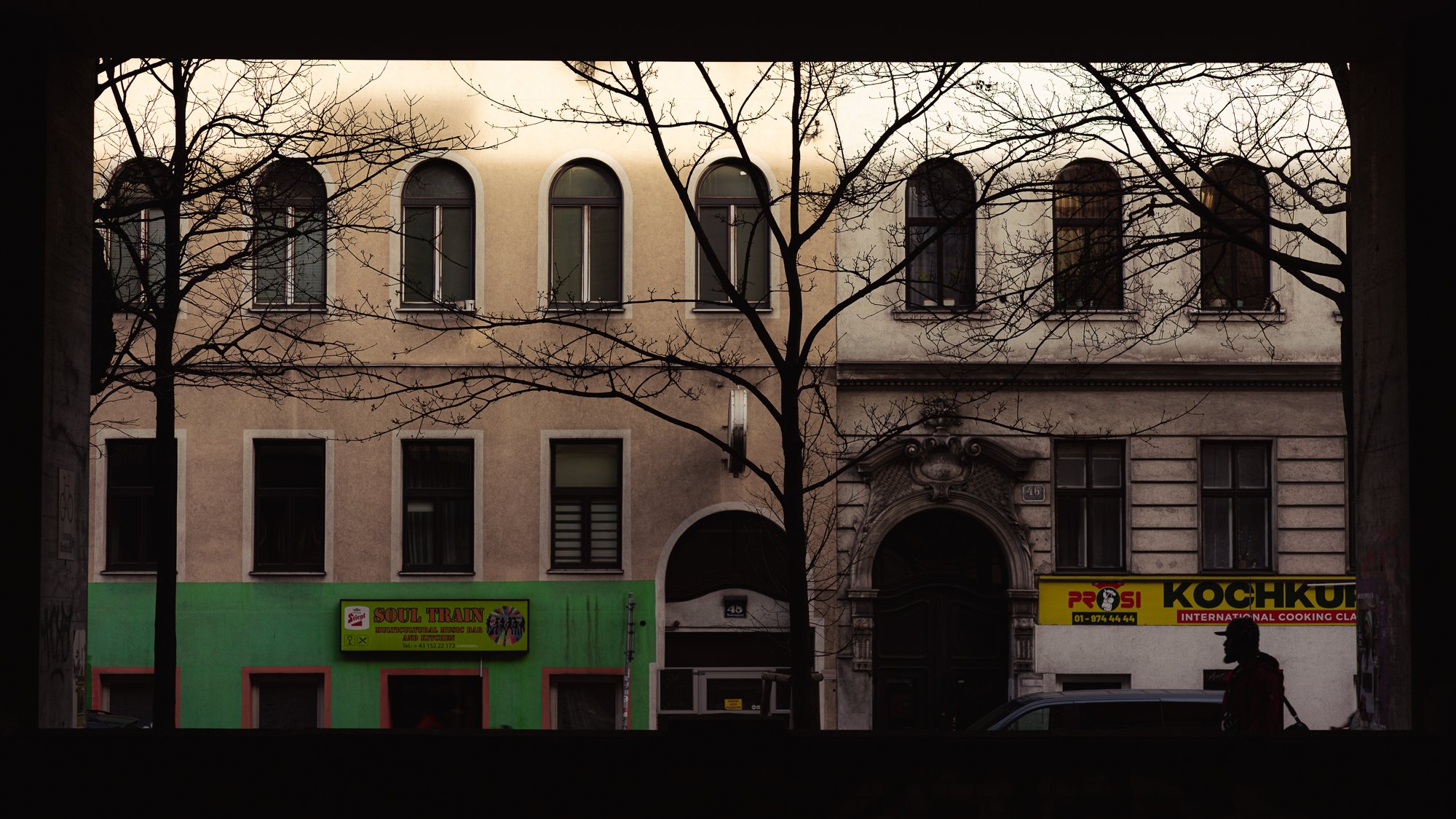 View of a building through a window with leafless trees in the foreground, and shop signs on the street below.