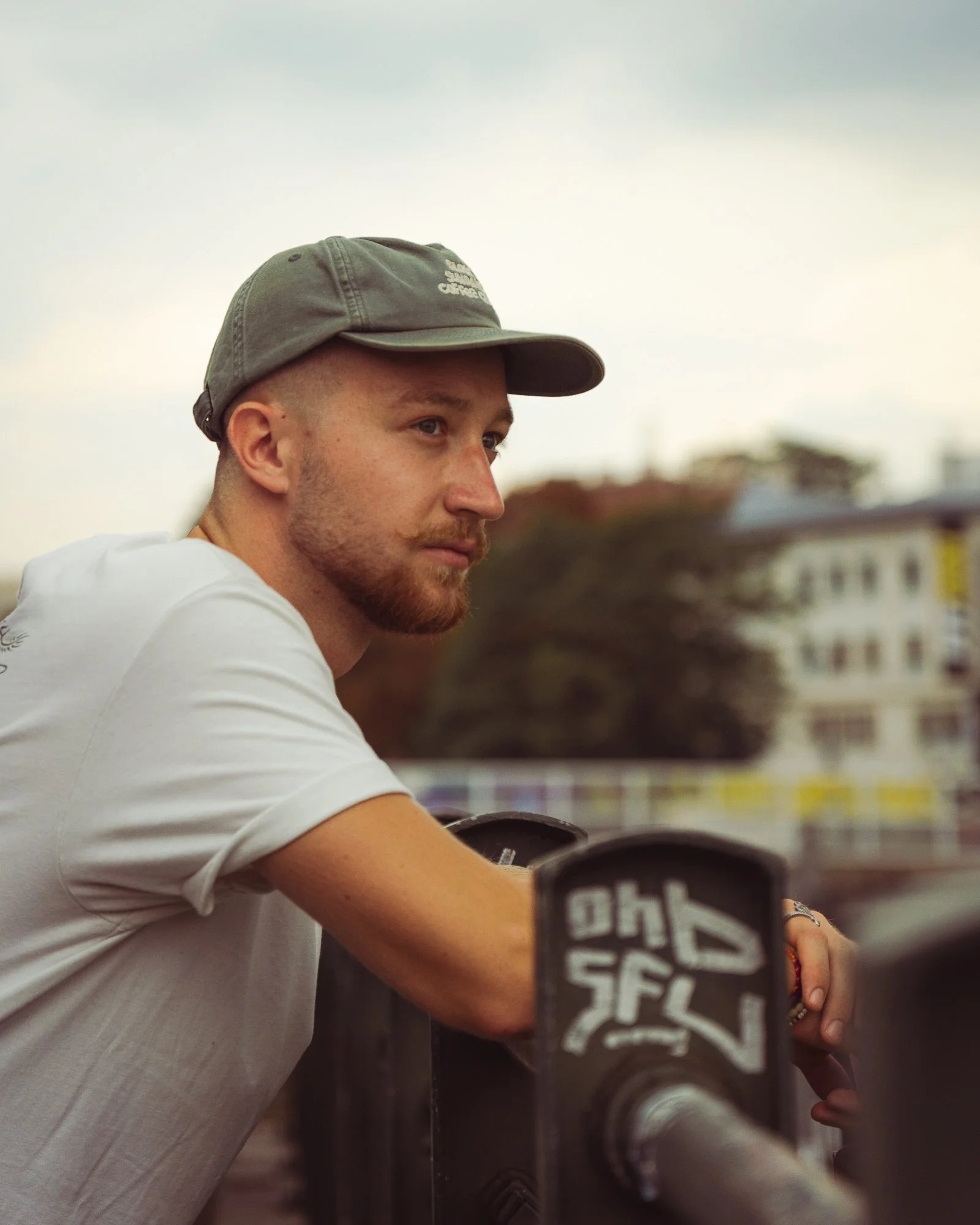 A young man with a beard and light skin wearing a gray baseball cap and white T-shirt, sitting outdoors with a blurred cityscape in the background.