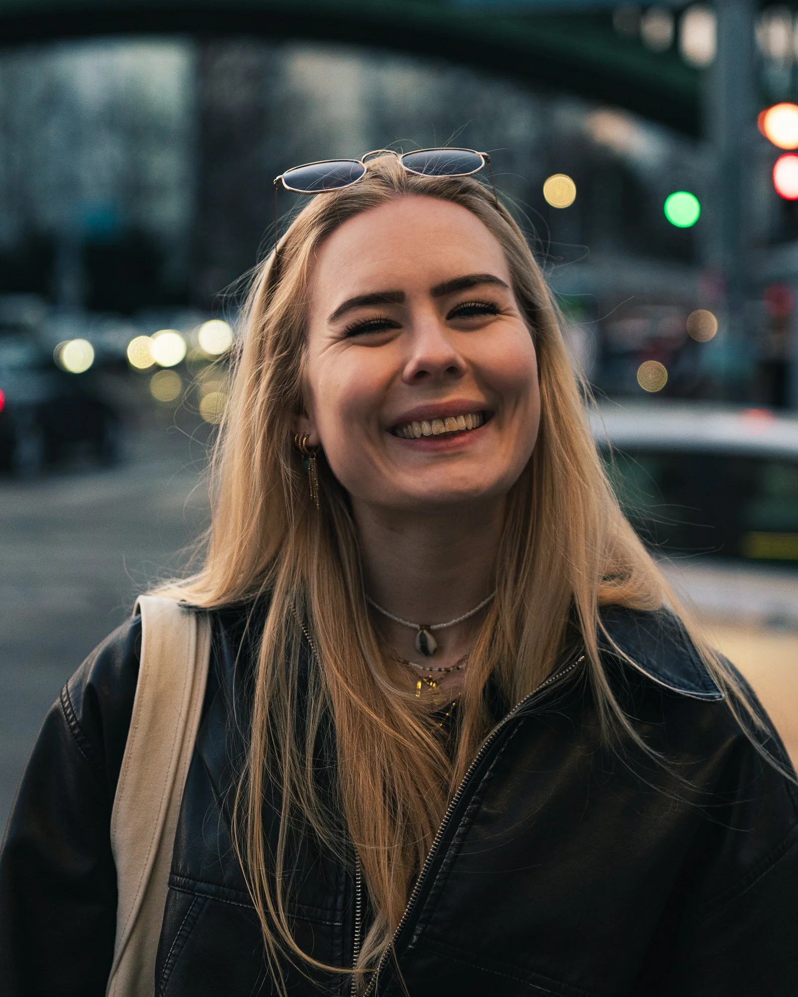 A smiling young woman with blonde hair, wearing sunglasses on her head, a black leather jacket, layered necklaces, and a beige shoulder bag, standing outdoors with blurred city traffic and lights in the background.