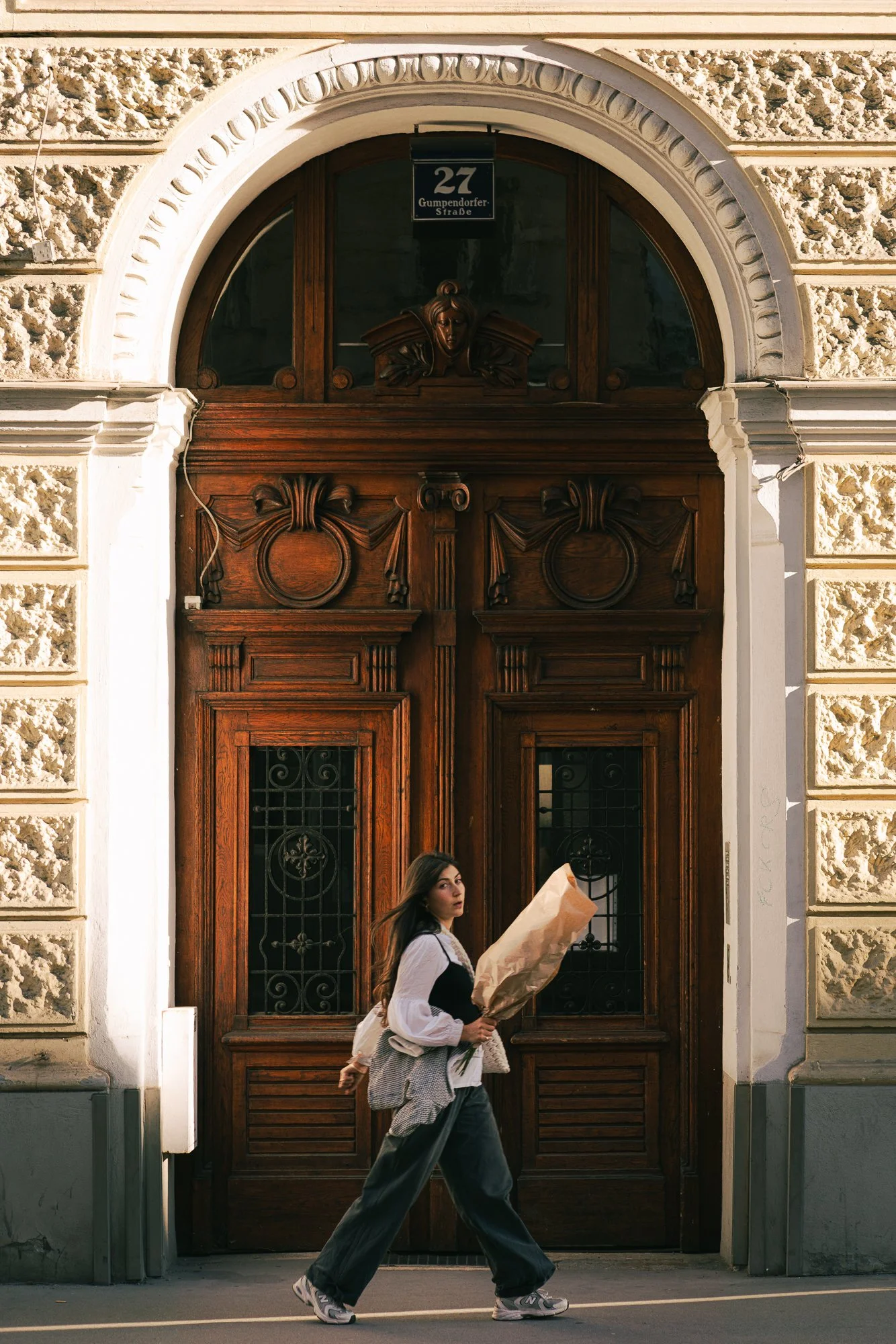 A woman walking in front of a large, ornate wooden door with an arched window above it, holding a bouquet wrapped in brown paper.