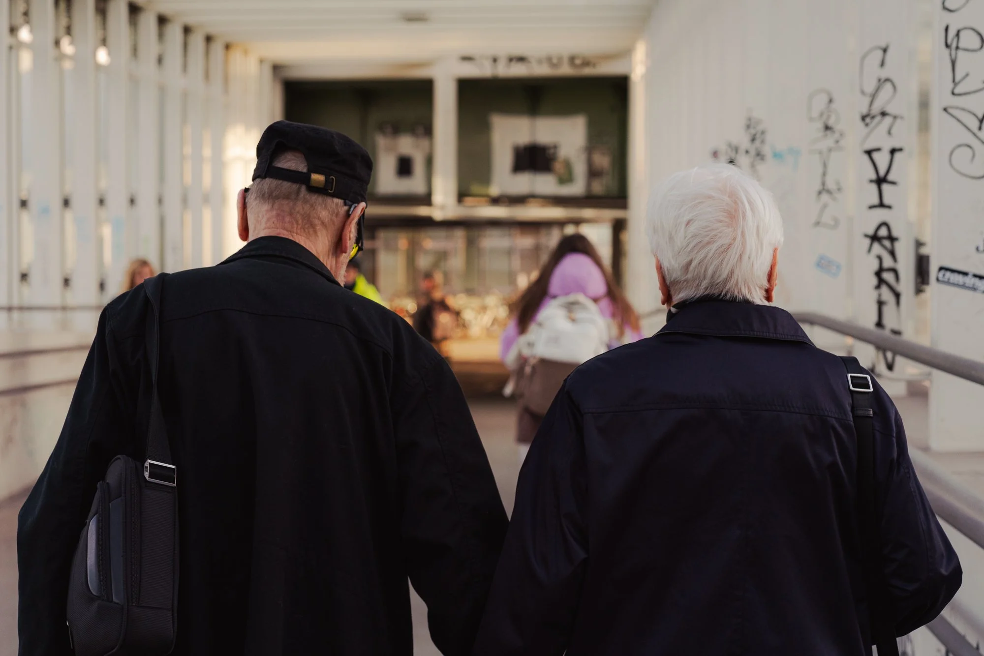 Two older men with gray and white hair, wearing dark clothing, are walking away from the camera in a covered pedestrian bridge with graffiti on their sides. The background shows young people walking ahead, some carrying backpacks.