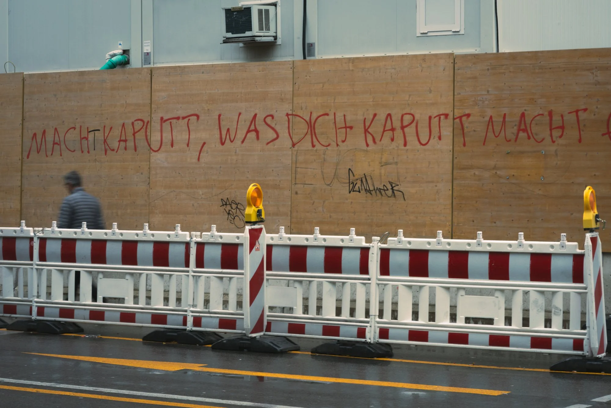 A construction site with a tall wooden barrier with red graffiti writing in a foreign language. There are traffic barriers and cones in front of the barrier, and a man in a striped jacket walking past. The background features the upper part of a buil