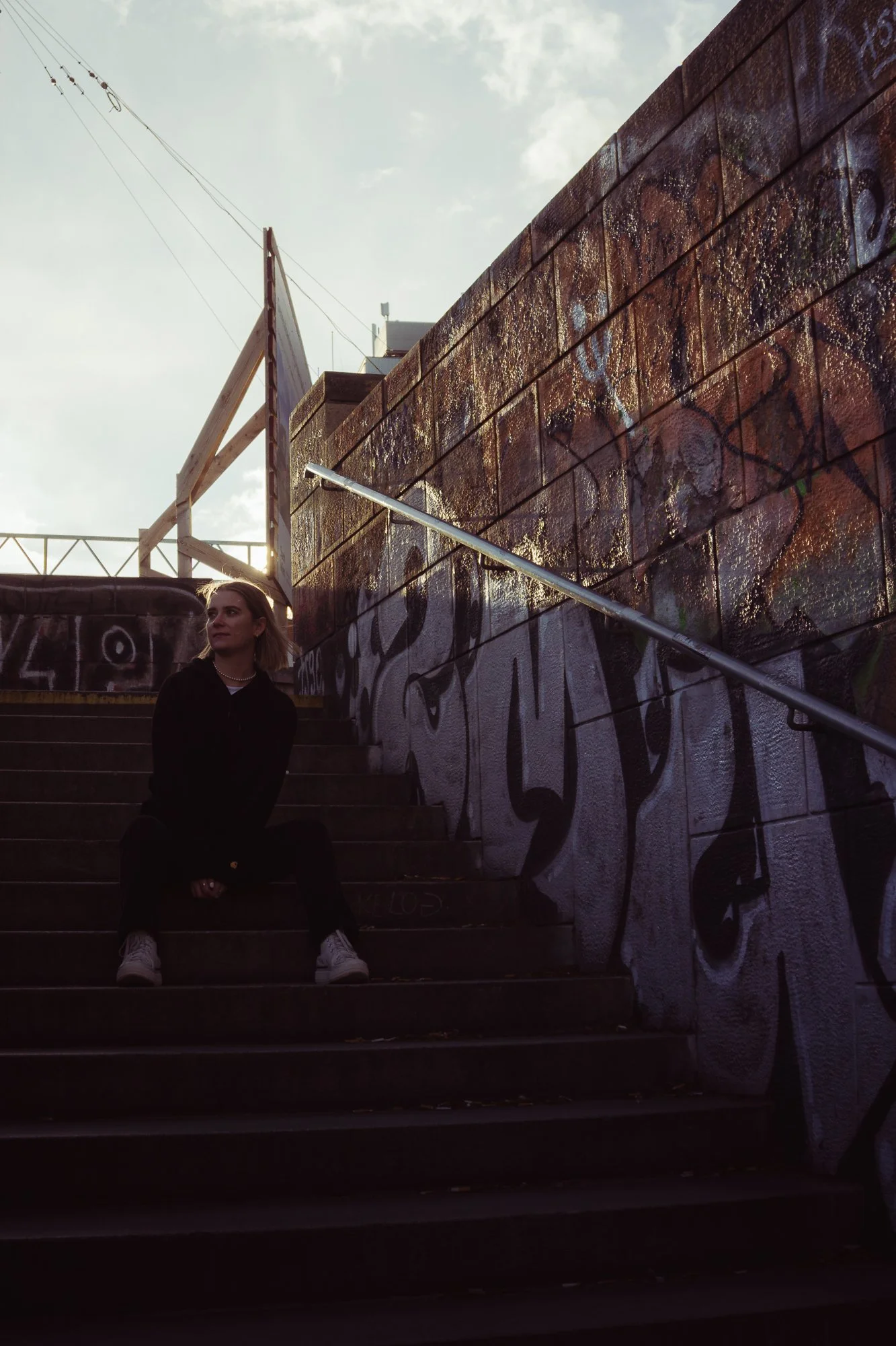 A woman in black clothing and white shoes sitting on outdoor steps at dusk, with graffiti-covered walls and urban setting in the background.
