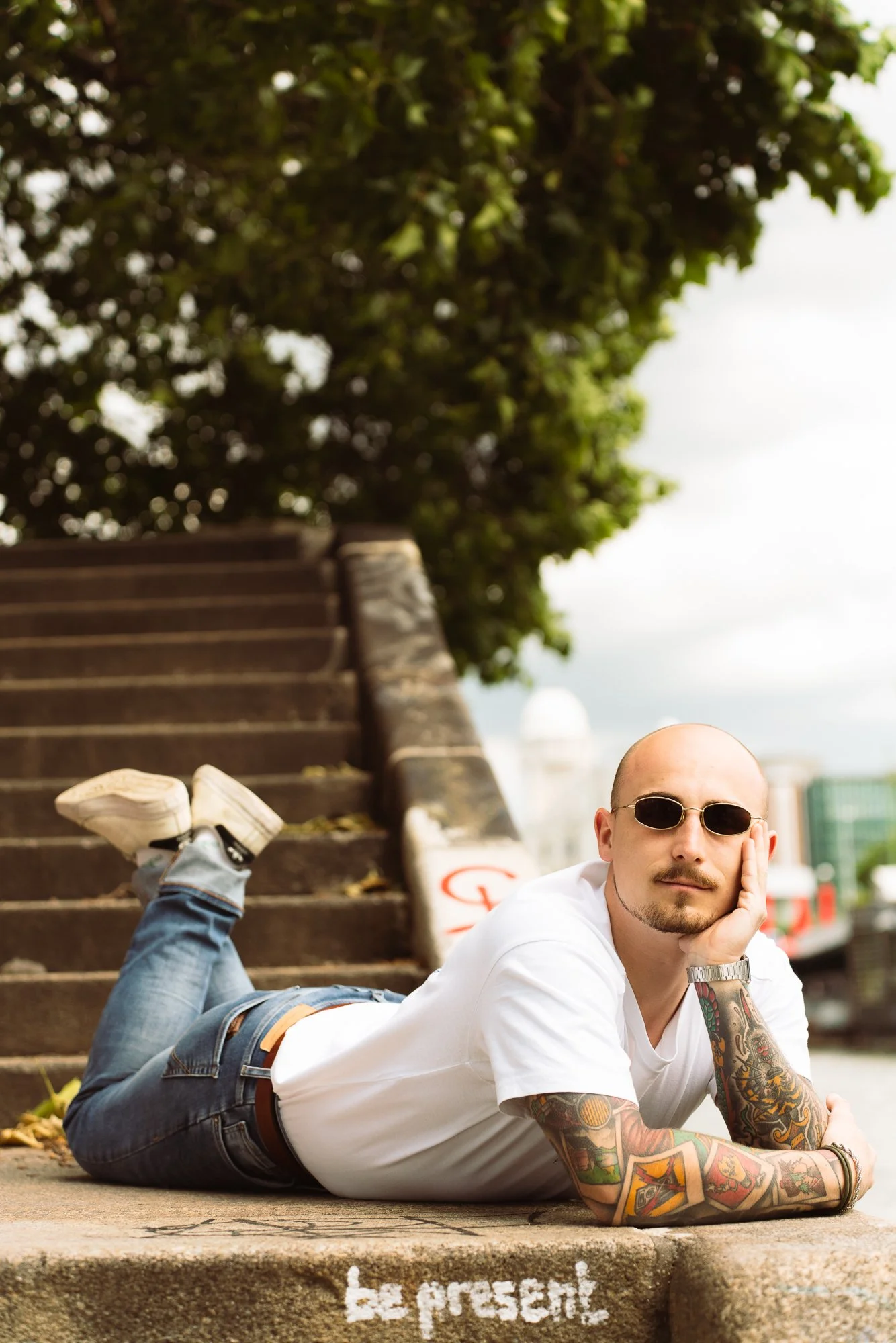A young man with tattoos, wearing sunglasses, lying on his side on a city sidewalk. He has his hand on his face and is looking into the distance. There are stairs and a tree in the background.