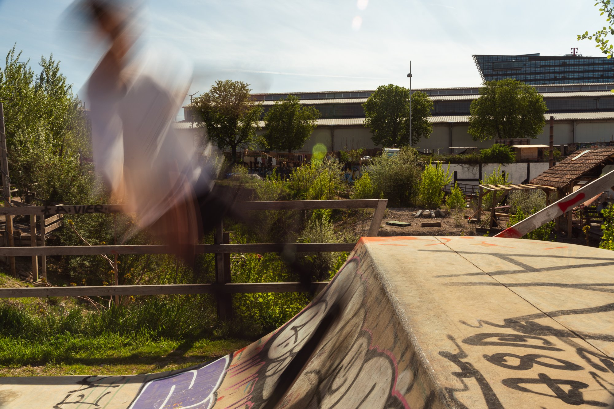 Blurred person in the foreground, skate ramp with graffiti, trees, industrial building, and modern glass building in the background on a sunny day.