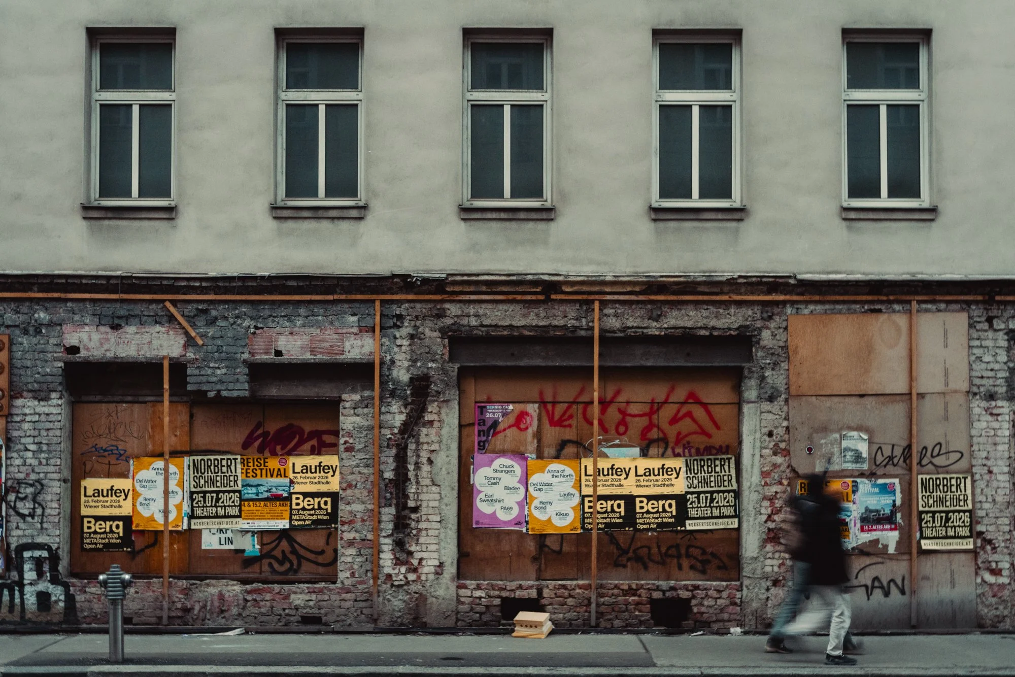 A partially renovated building with four windows on an upper level and a boarded-up lower level covered in graffiti and posters; a person walking past in the foreground.