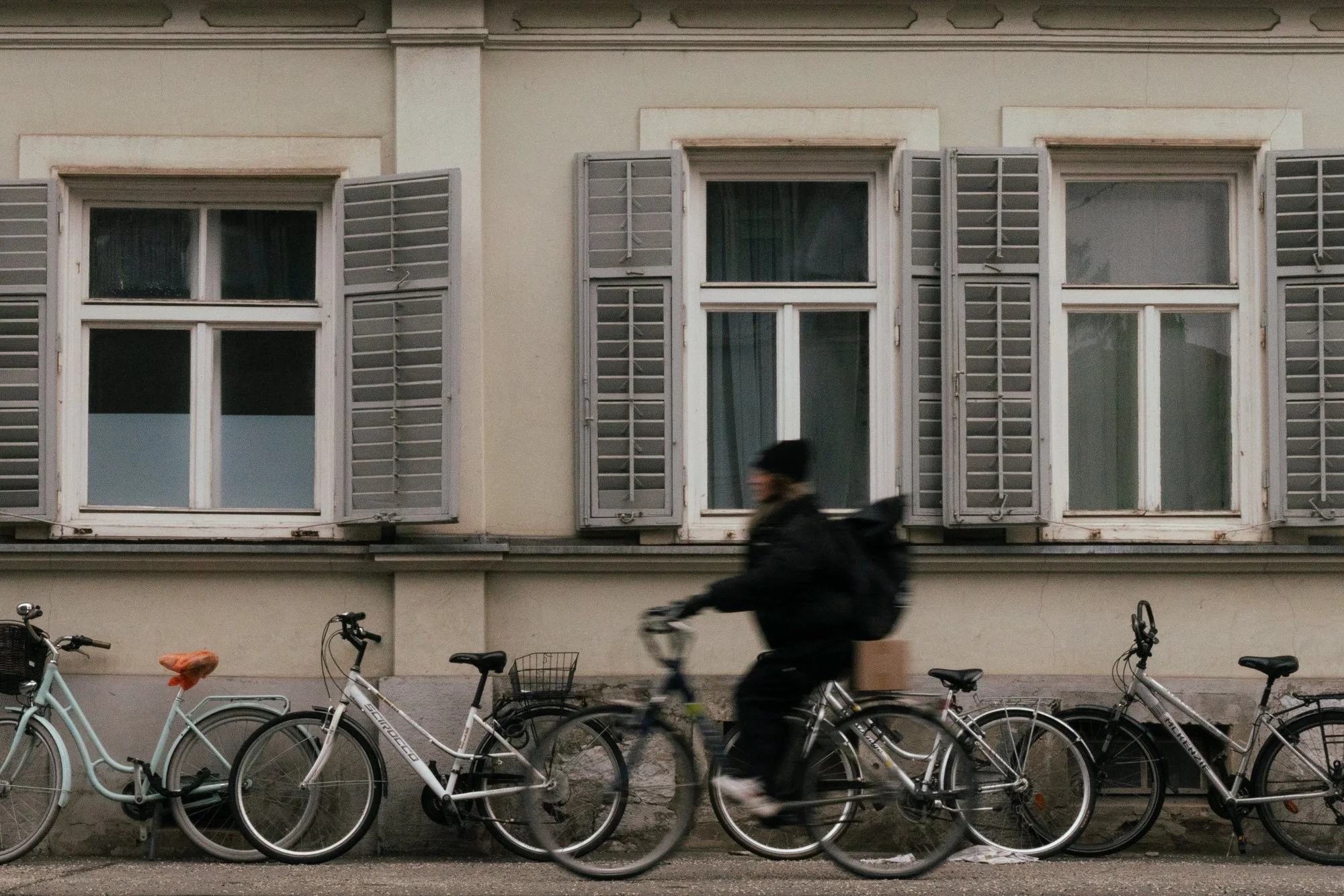 A person riding a bicycle past a row of parked bicycles in front of a building with three windows and gray shutters.
