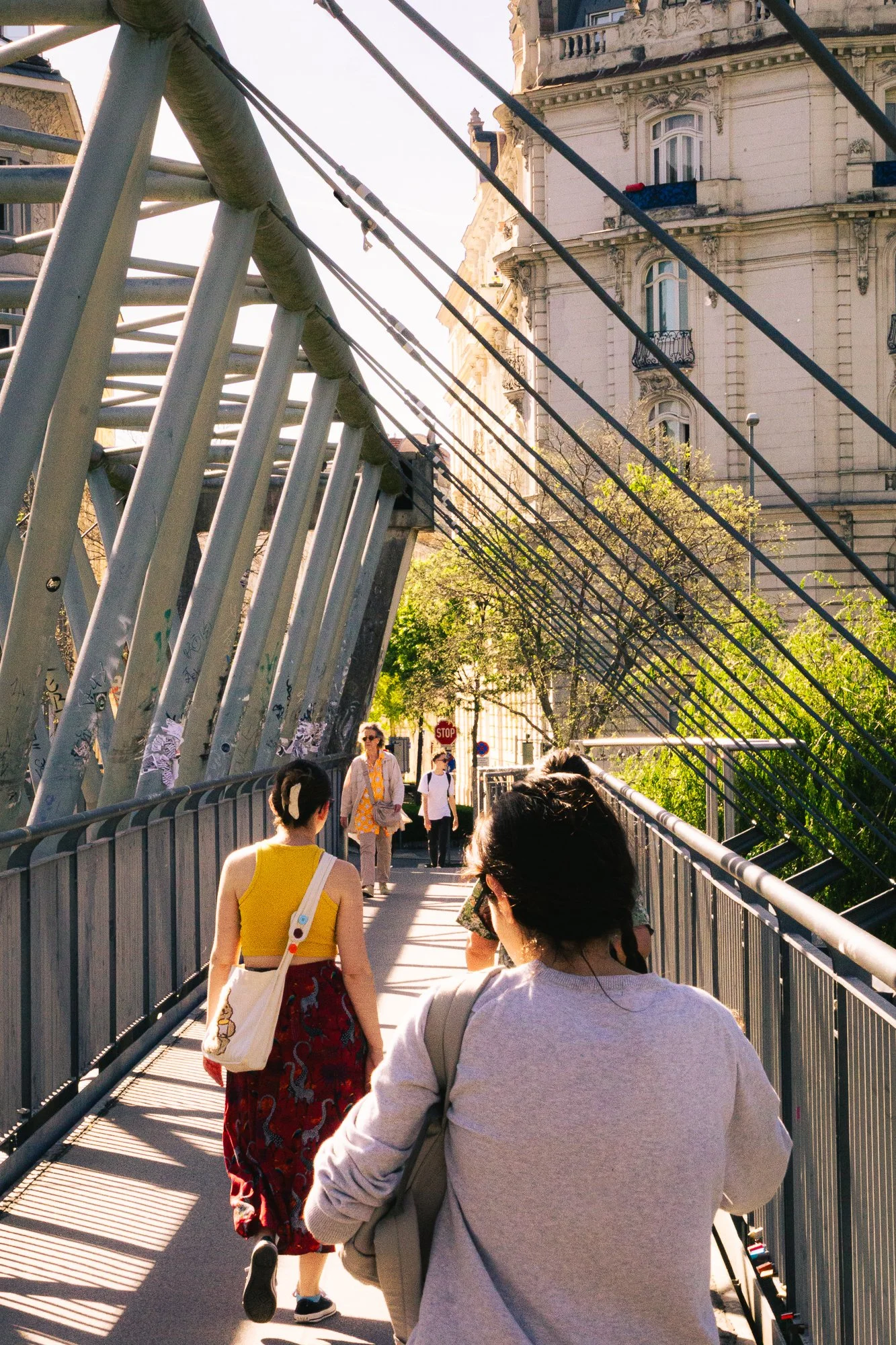 People walking across a modern pedestrian bridge with metal railings and cables, with old European-style buildings in the background.