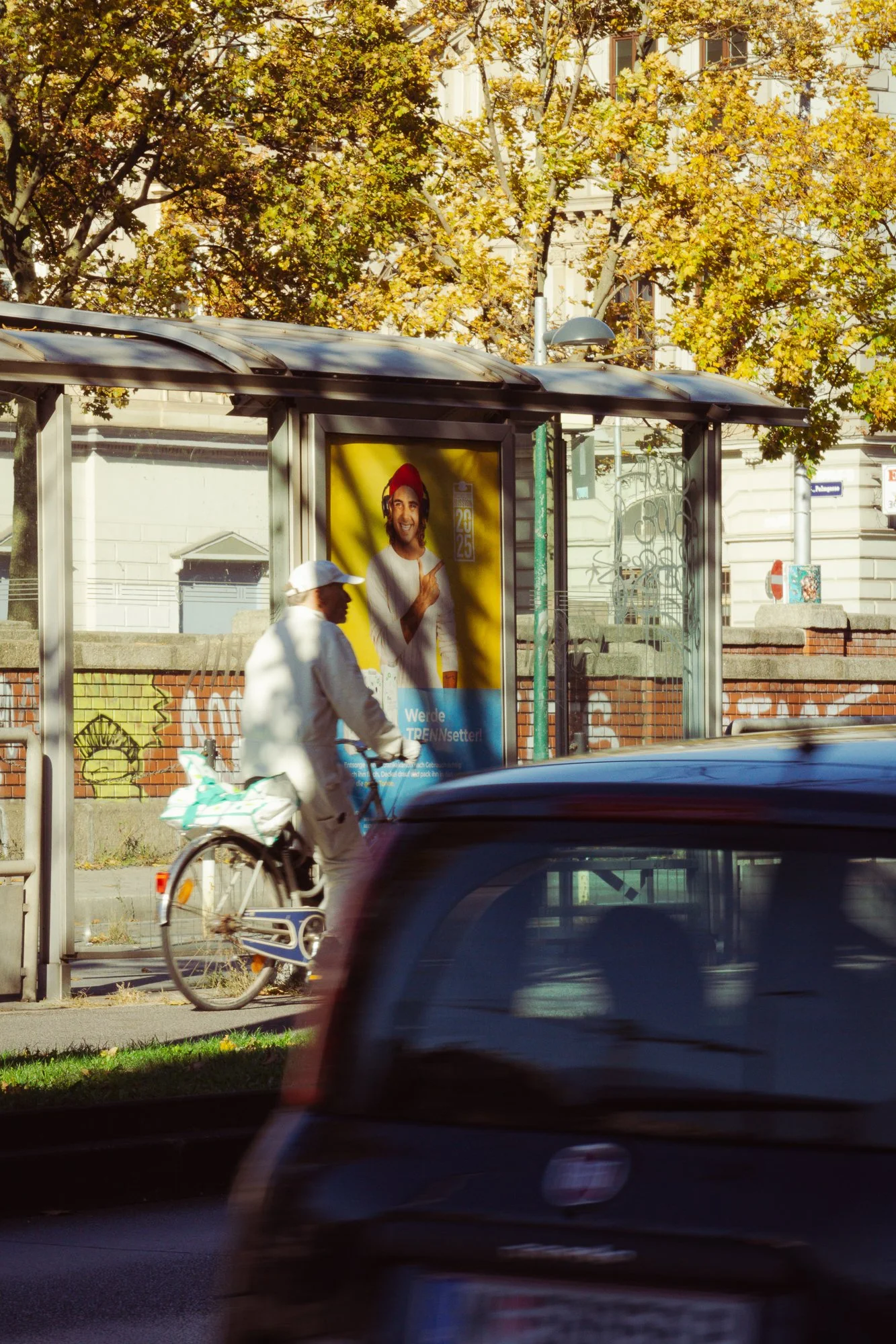 A cyclist wearing white clothing and a white cap is riding a bicycle past a bus shelter with an advertisement of a smiling man with red hair and a beard, in an urban area with trees and buildings in the background.
