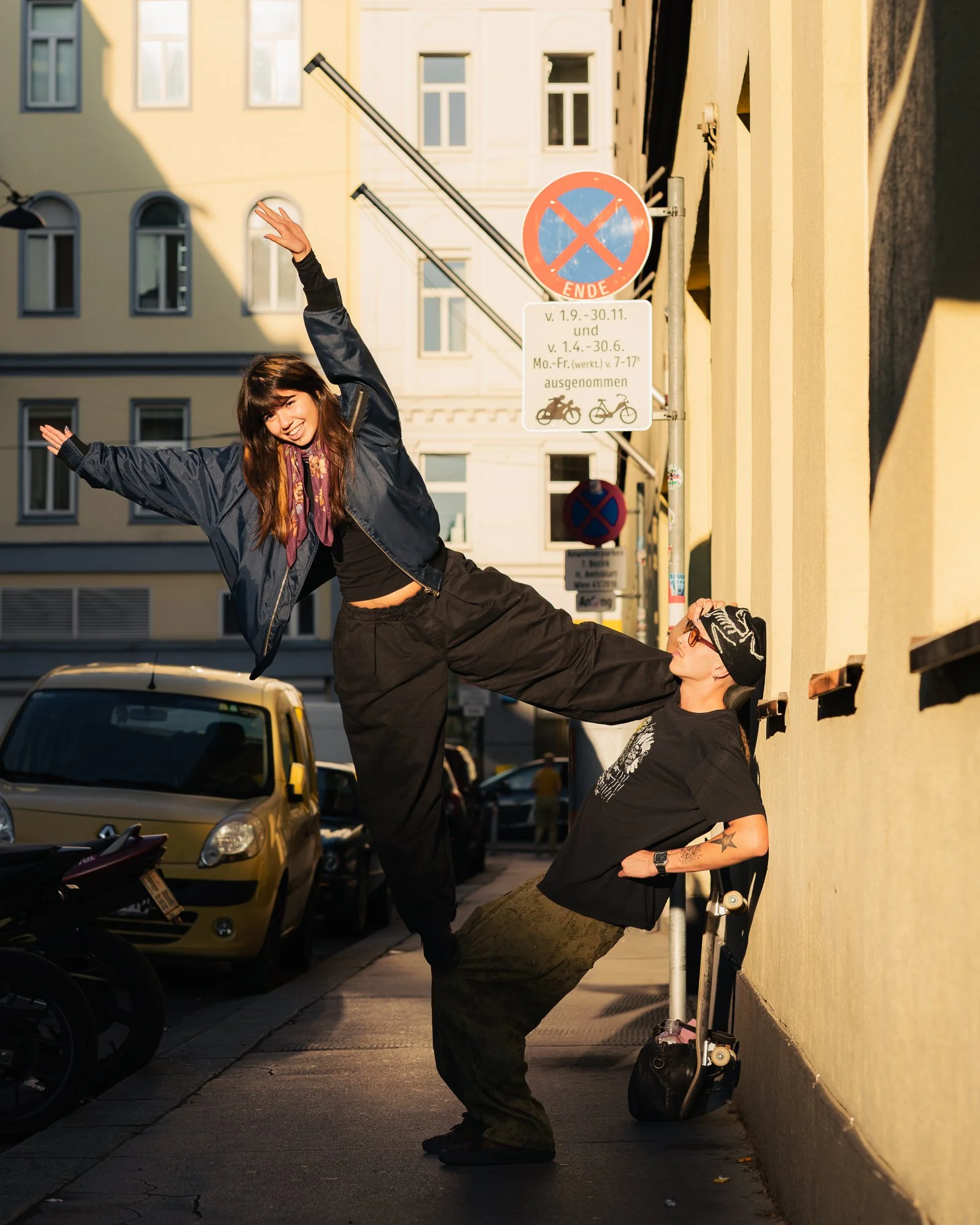 Two people, a woman and a man, are performing skateboard tricks on a city sidewalk, with buildings and cars in the background. The woman is smiling and raising her hand, while the man is holding onto the wall.