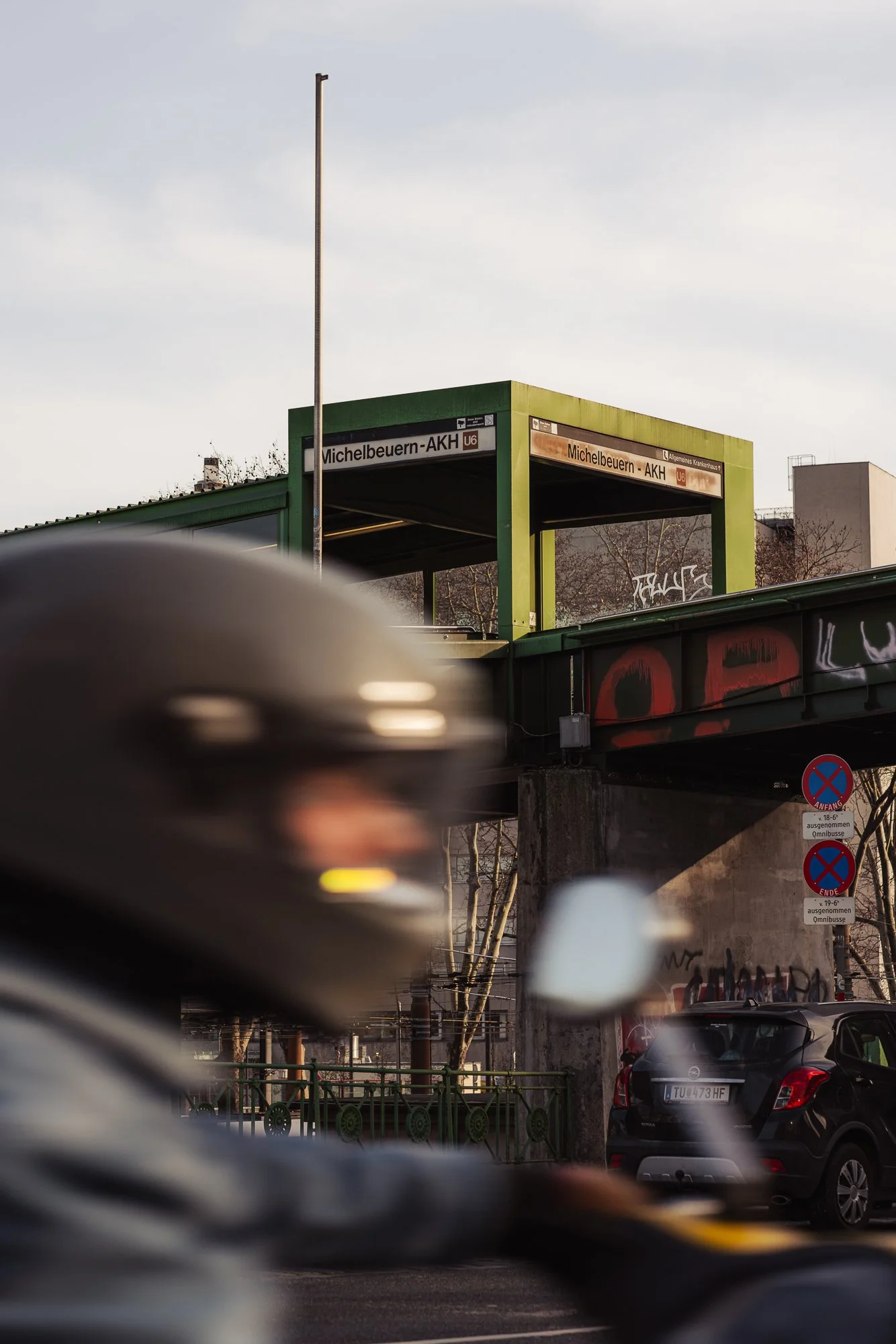 View of a train station entrance labeled "Michelbeuern-AKH" in Vienna, Austria, with surrounding cars and street signs, captured through an out-of-focus foreground of cars.