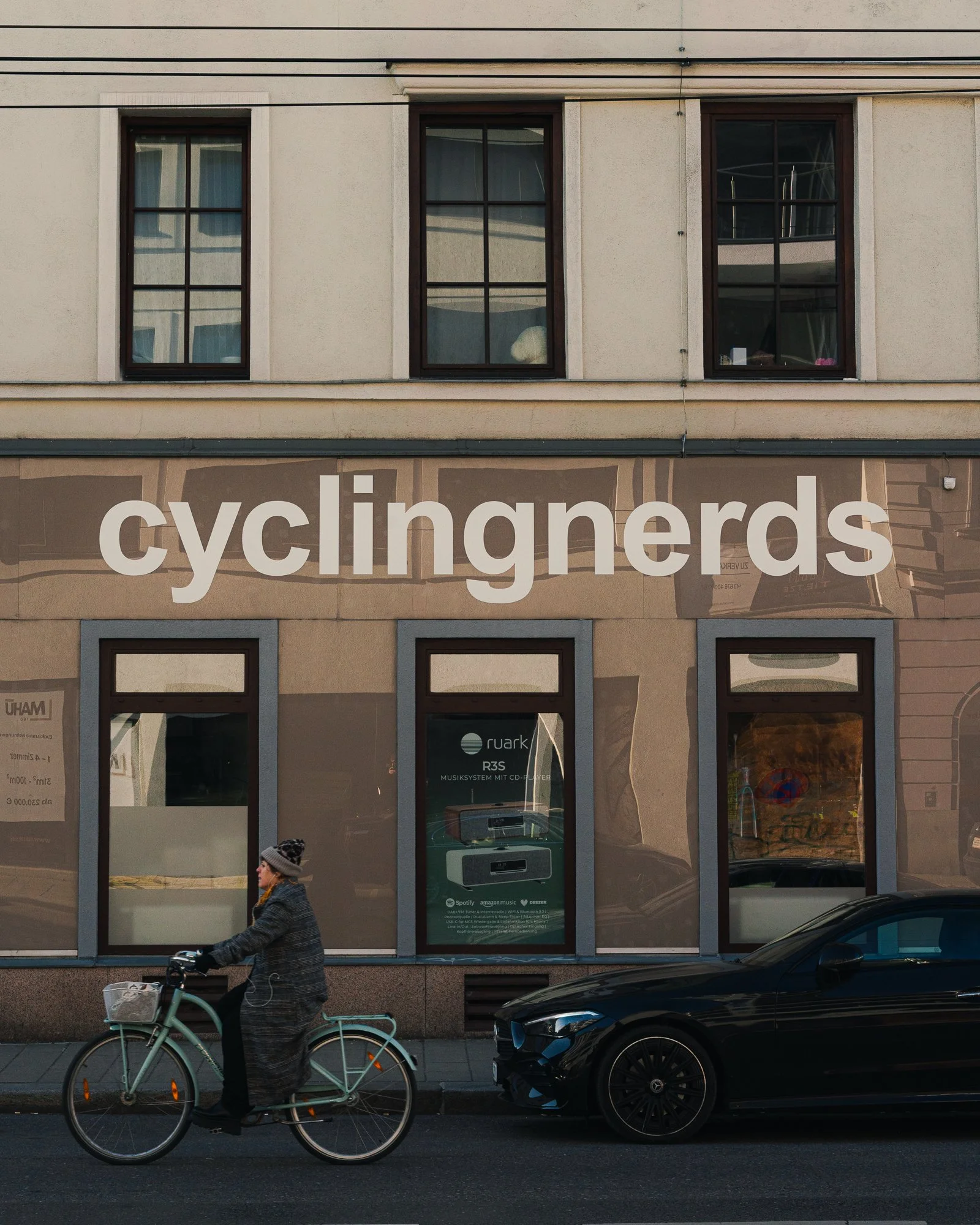 A woman wearing a gray coat, hat, and glasses is riding a light blue bicycle with a front basket on the street. In the background, there is a building with two stories. The building has three large windows on the first floor and three windows on the 