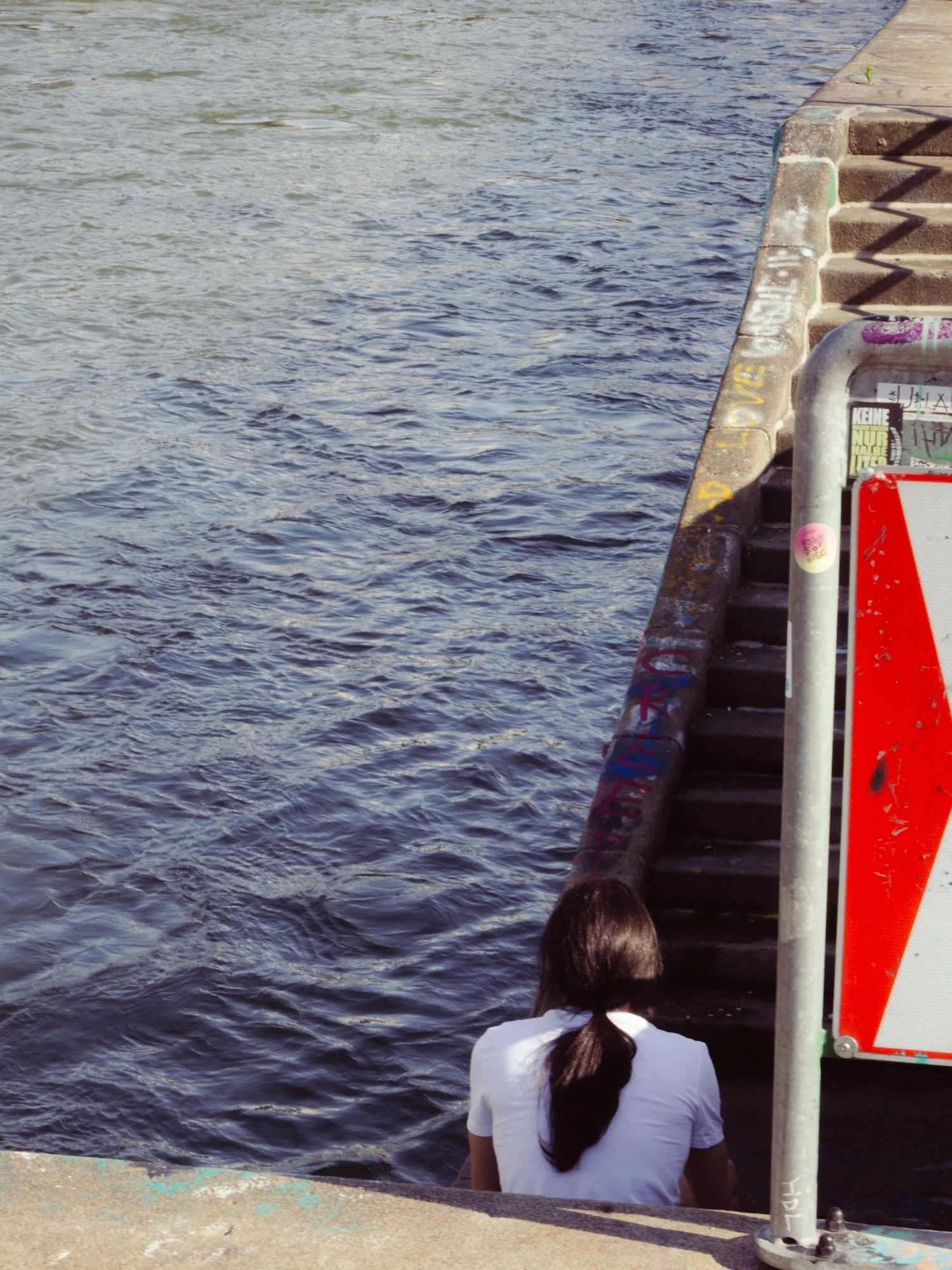 A woman with long dark hair tied in a ponytail, wearing a white shirt, sitting on the edge of a dock next to water with steps leading down to it. There are graffiti and stickers on the side of the steps and railing.