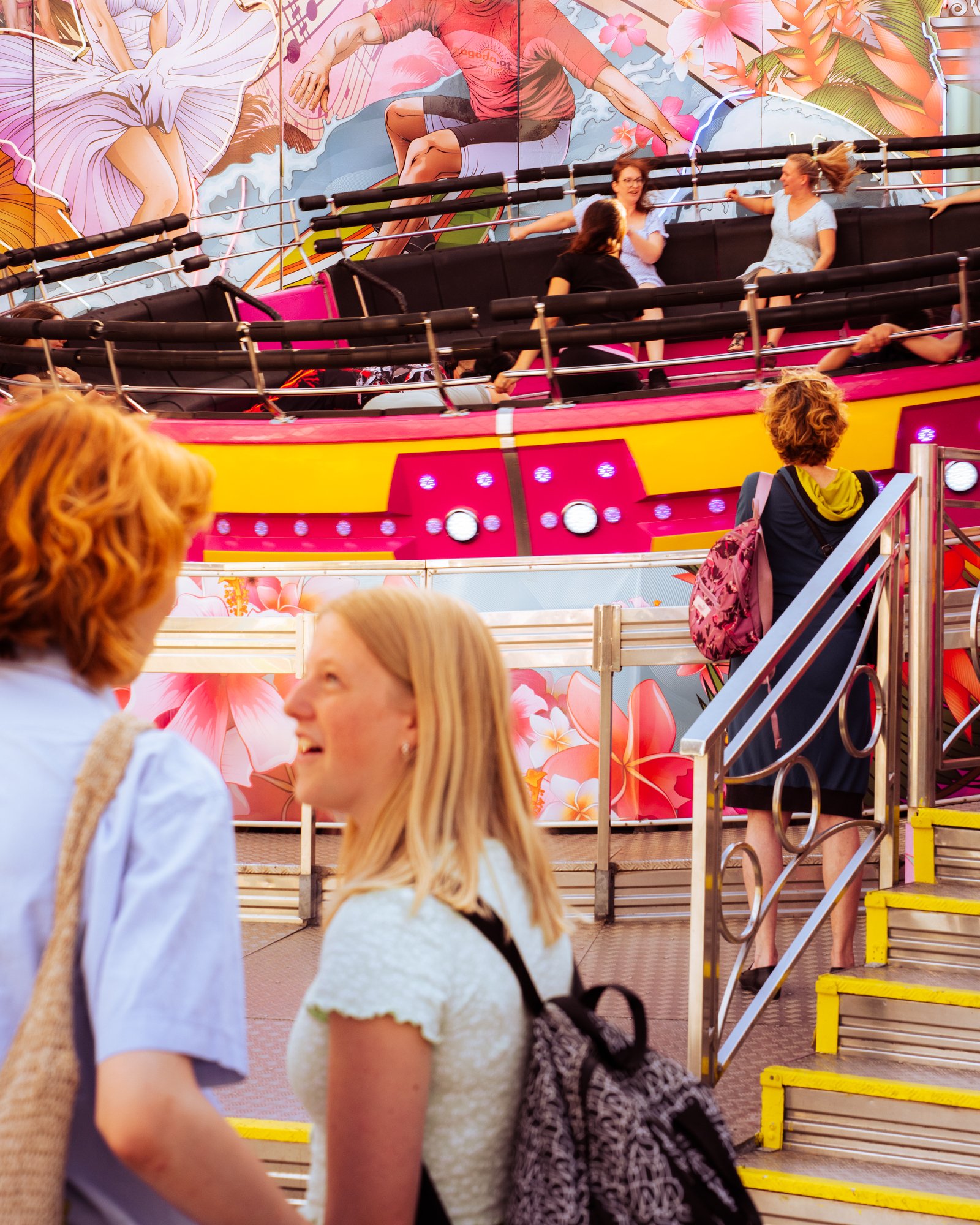 People riding an amusement park roller coaster with vibrant floral and ocean-themed artwork in the background.