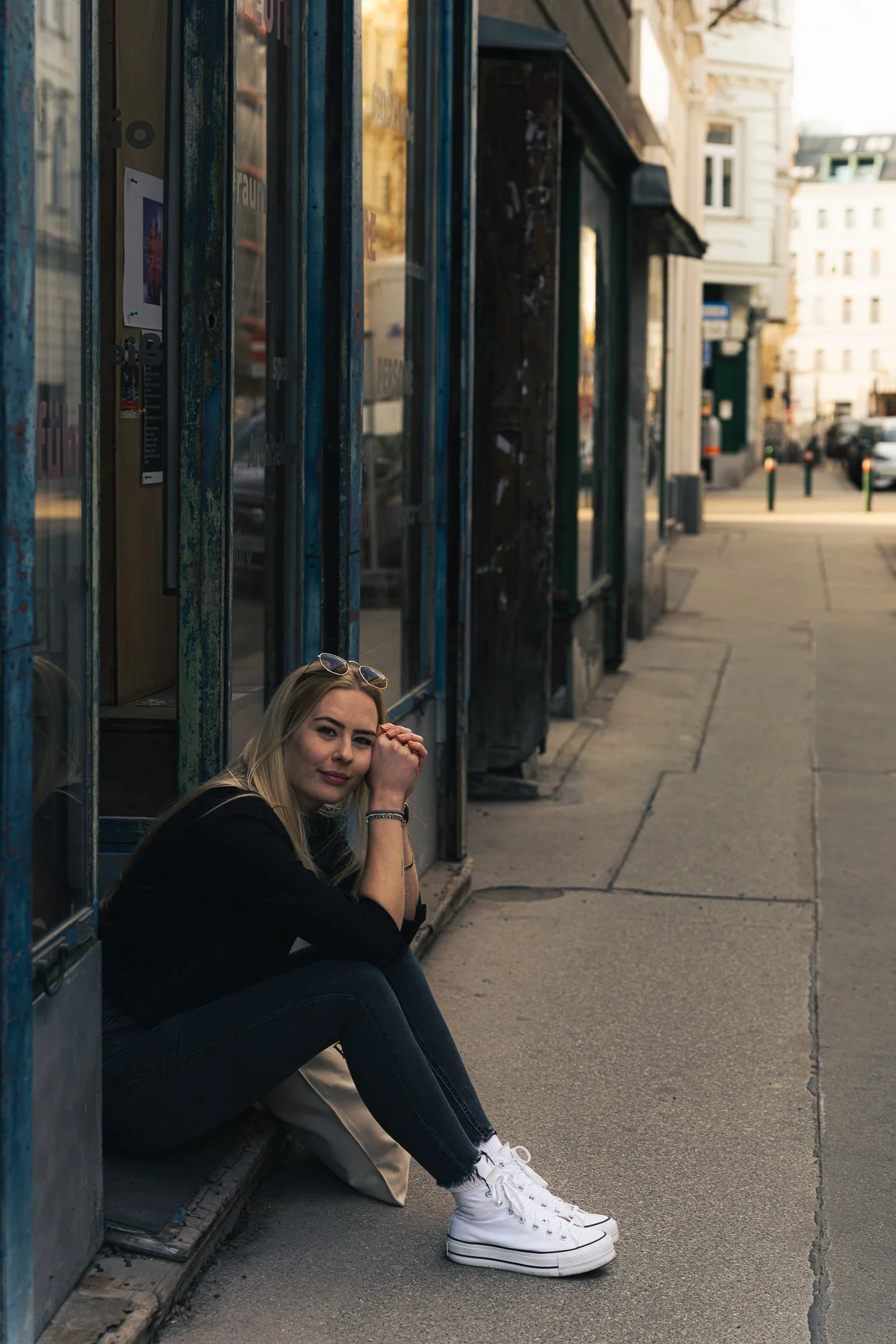 A young woman with blonde hair and sunglasses on her head sitting on the sidewalk beside a row of glass-paneled phone booths in an urban area during late afternoon or early evening, with buildings and parked cars visible in the background.