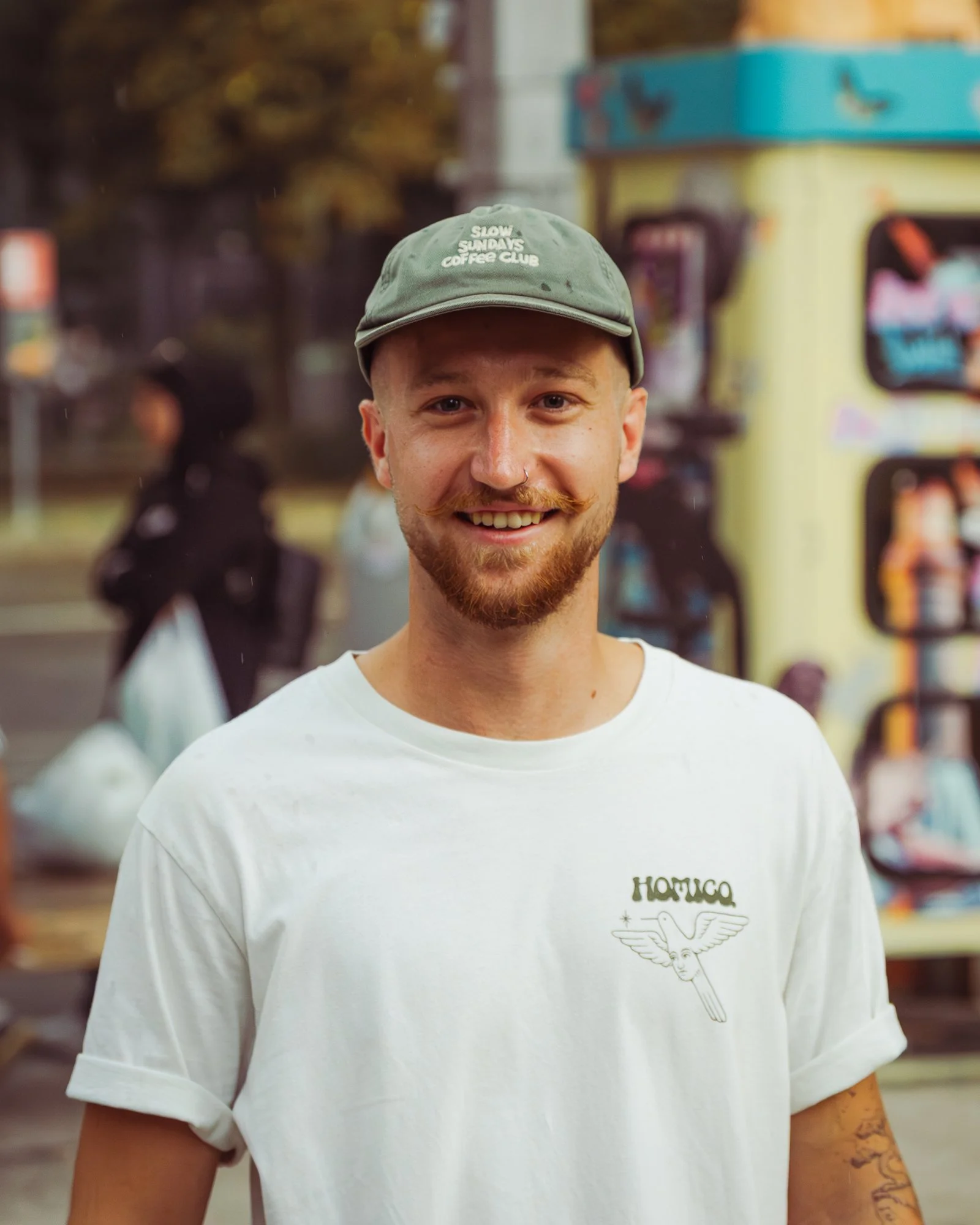 Smiling young man with a nose piercing, mustache, and beard wearing a gray cap and a white T-shirt with a graphic design, standing in front of a colorful background with people and vending machines.