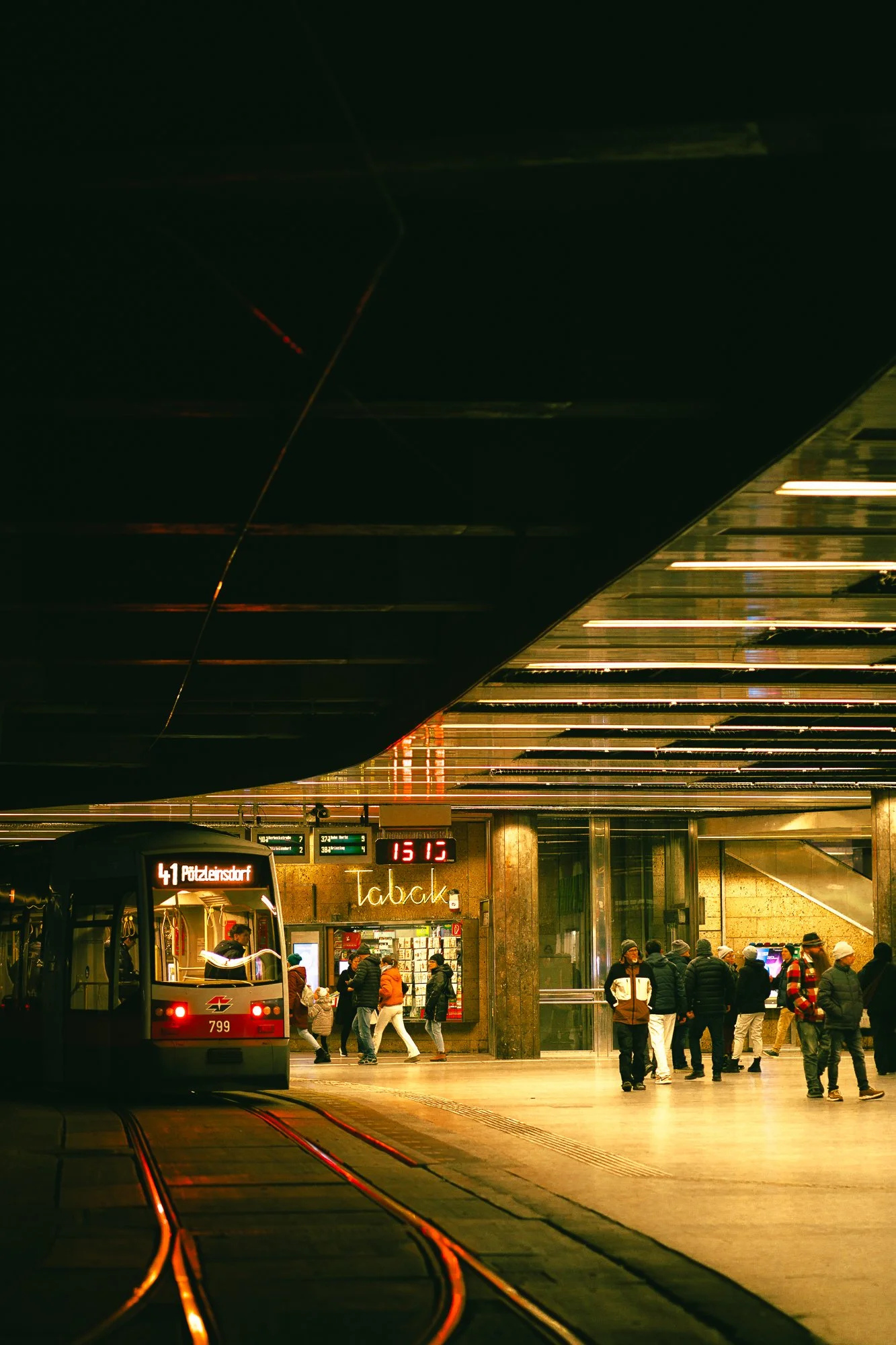 A busy train station with a train on the tracks, travelers walking, and a restaurant named 'Tabak' in the background. The ceiling features lighting fixtures, and a digital clock reads 15:12.
