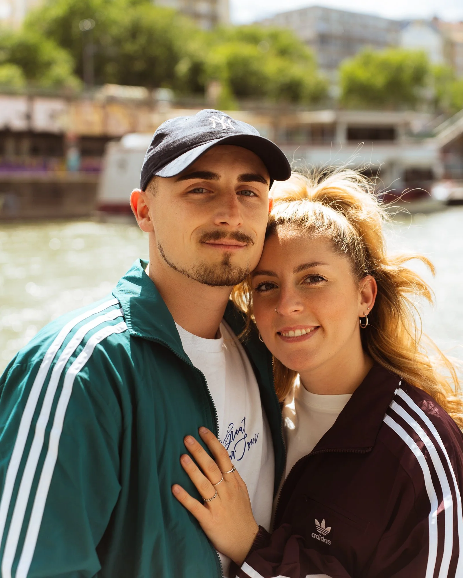 Close-up of a young man and woman in an outdoor setting, smiling and leaning close to each other, with a waterfront and greenery in the background.