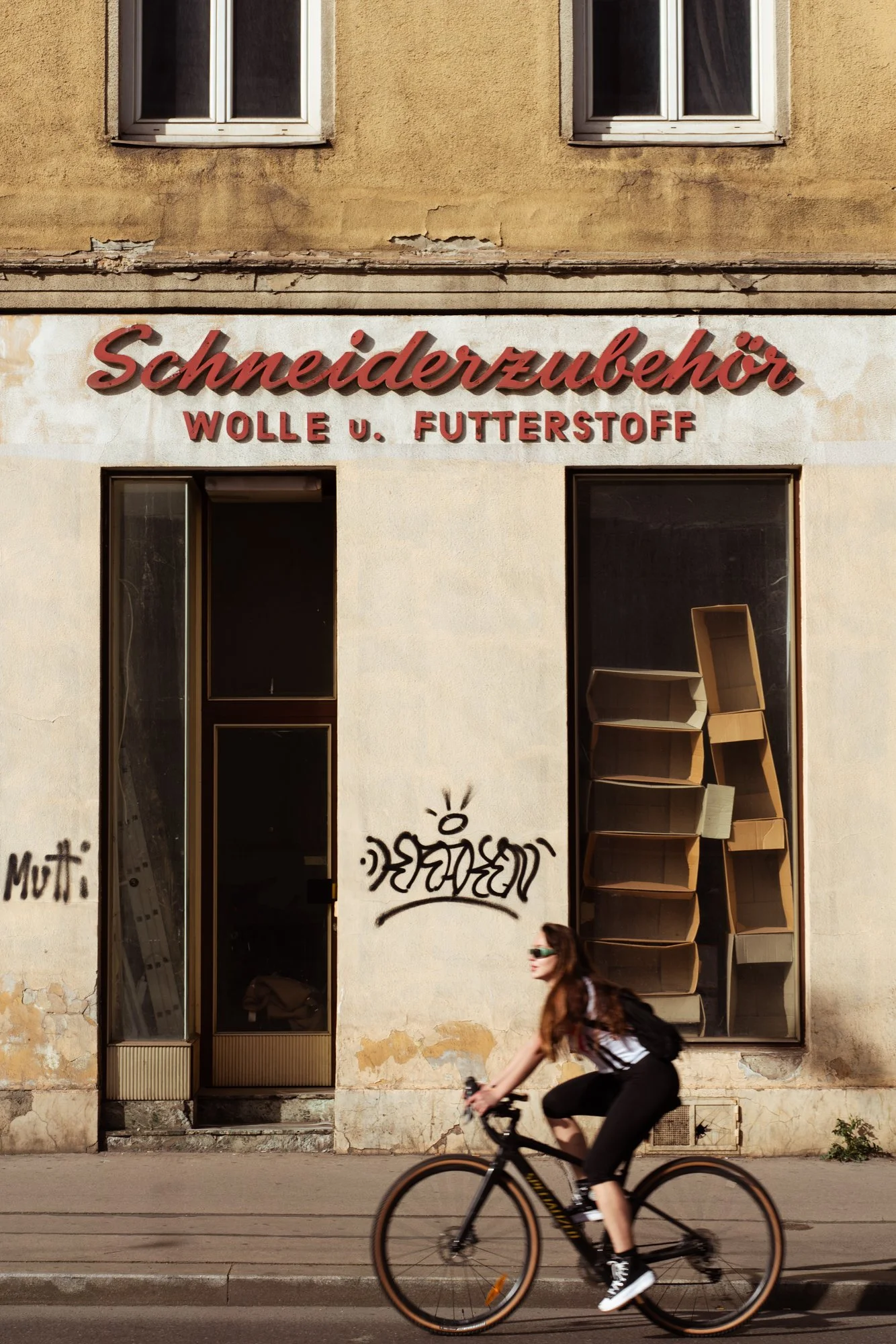 A woman riding a bicycle in front of a building with graffiti and a sign that reads 'Schneiderzuber Wolle u. Futterstoff'.