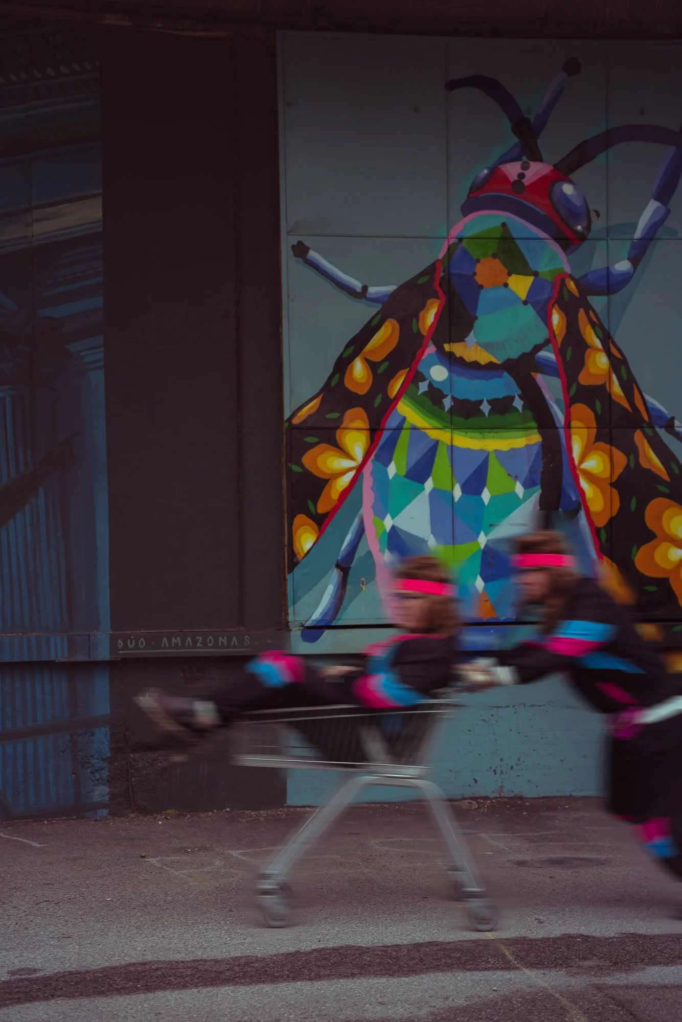 A mural of a colorful insect painted on a wall, with motion-blurred cyclists passing in front of it.