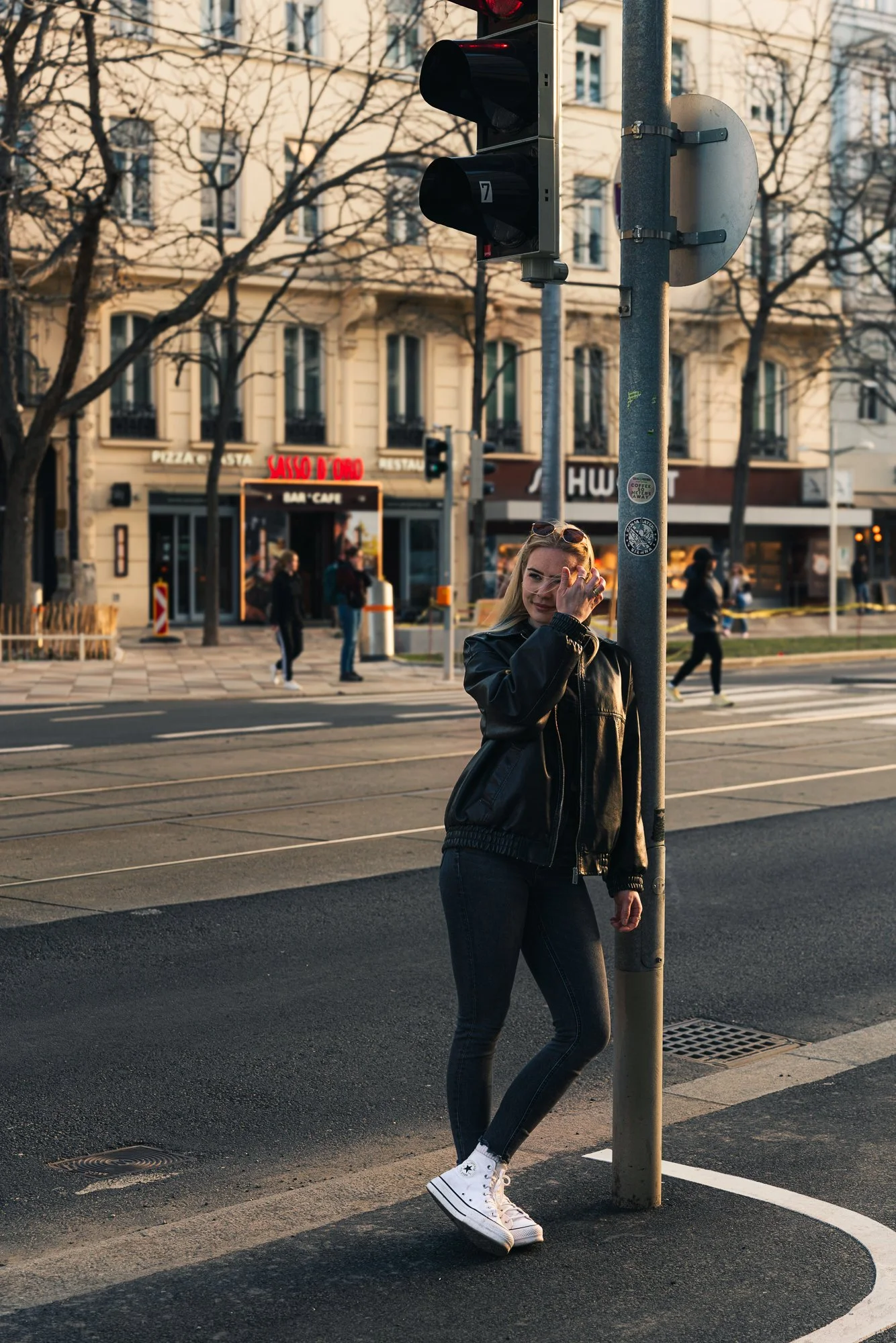 A woman standing near a streetlight on a city street, adjusting her sunglasses. She is wearing a black leather jacket, dark jeans, and white high-top sneakers. In the background, there are buildings, trees without leaves, and pedestrians passing by.