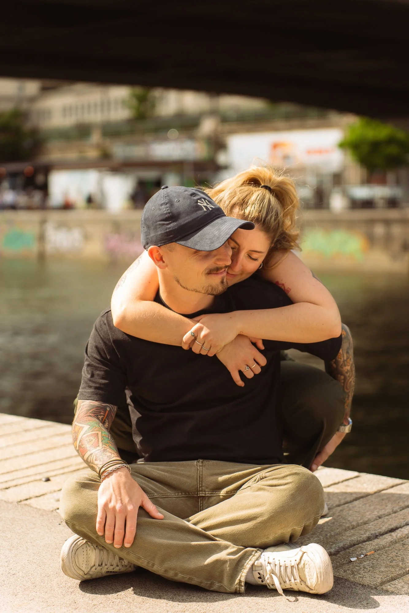 A couple sitting on a dock near water, with the woman hugging the man, both smiling and leaning their heads together.