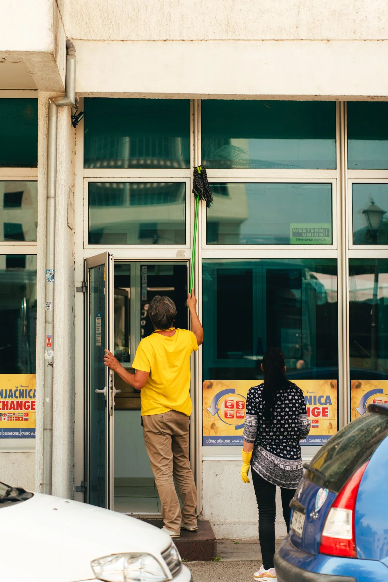 A person in yellow holding a mop and cleaning the glass door of a currency exchange office, with two people waiting outside.