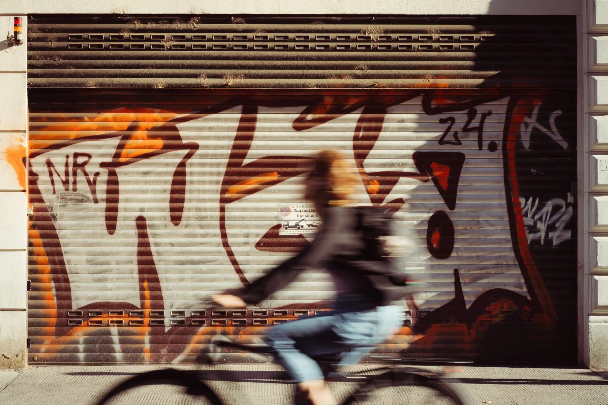 A person riding a bicycle past a graffiti-covered metal shutter door on a city street.