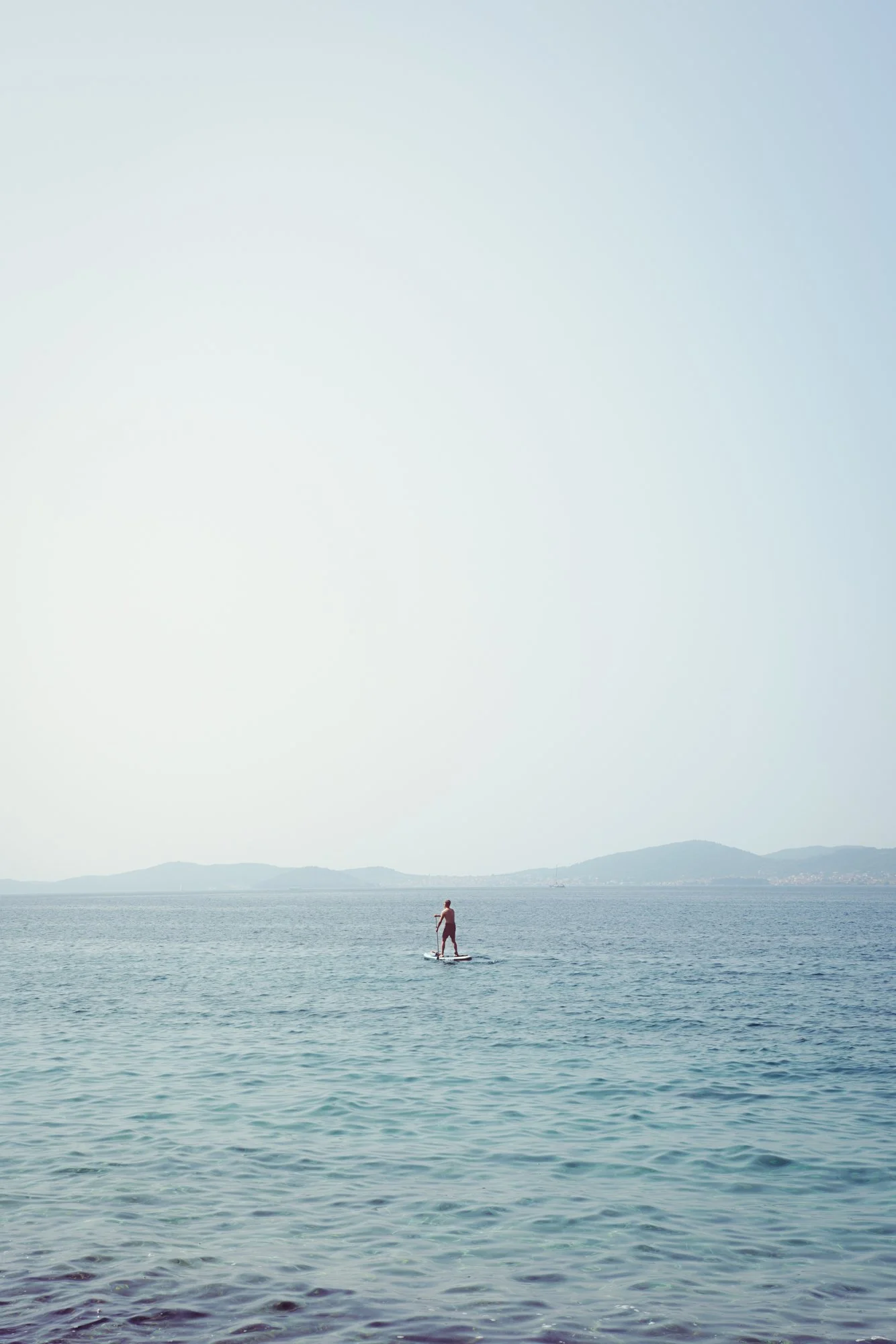 A person paddleboarding on a calm body of water with mountains in the background, under a pale sky.