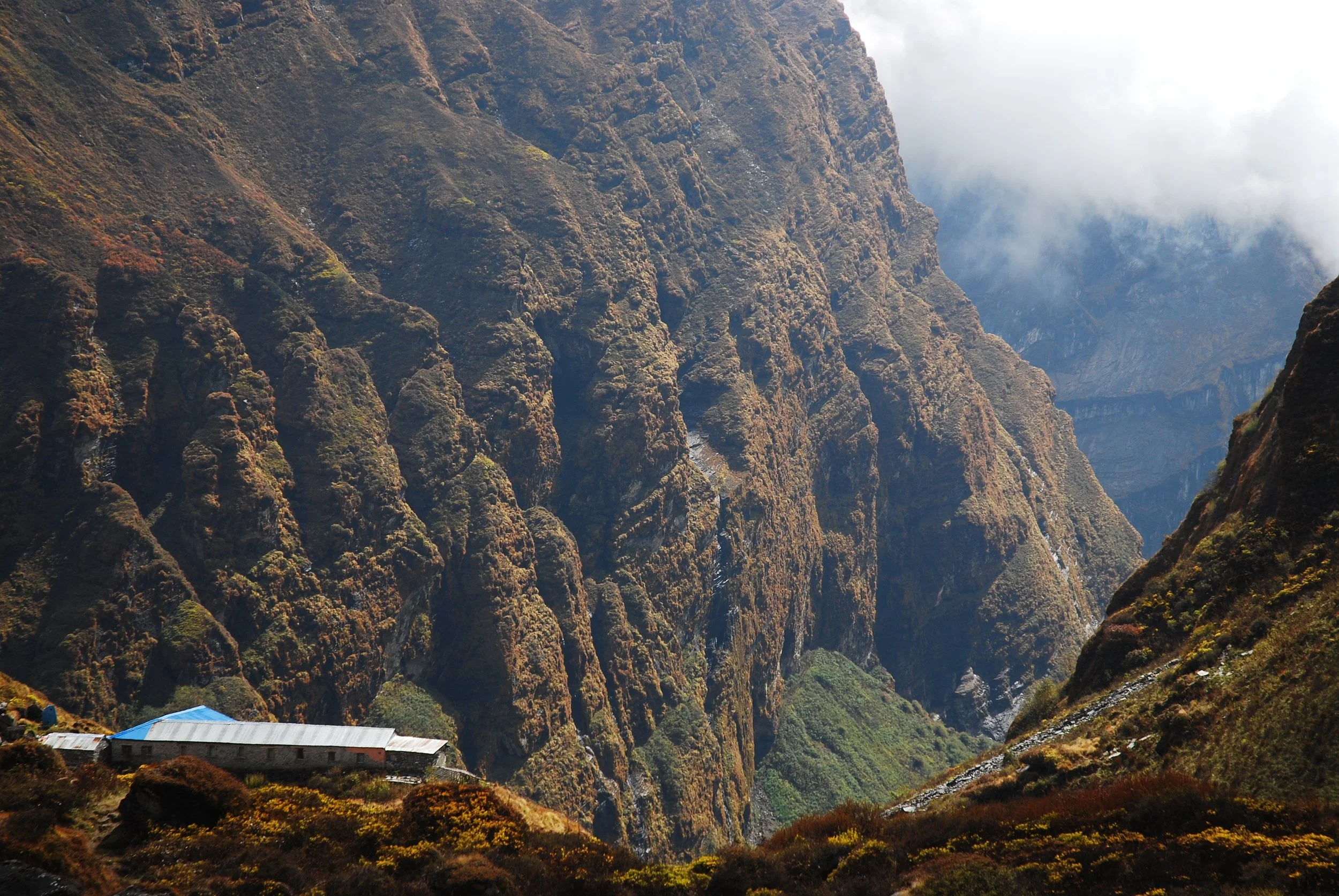 A mountain landscape with rugged, steep cliffs covered in green and brown vegetation. There is a small building with a blue roof situated in the lower left corner, with clouds or fog partially obscuring the distant peaks.