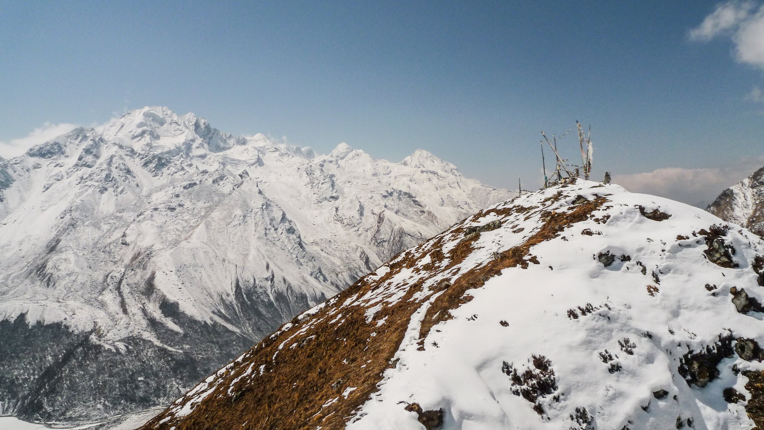 Snow-covered mountain range with a ridge in the foreground containing prayer flags and wooden poles, under a clear blue sky, in Langtang National Park.