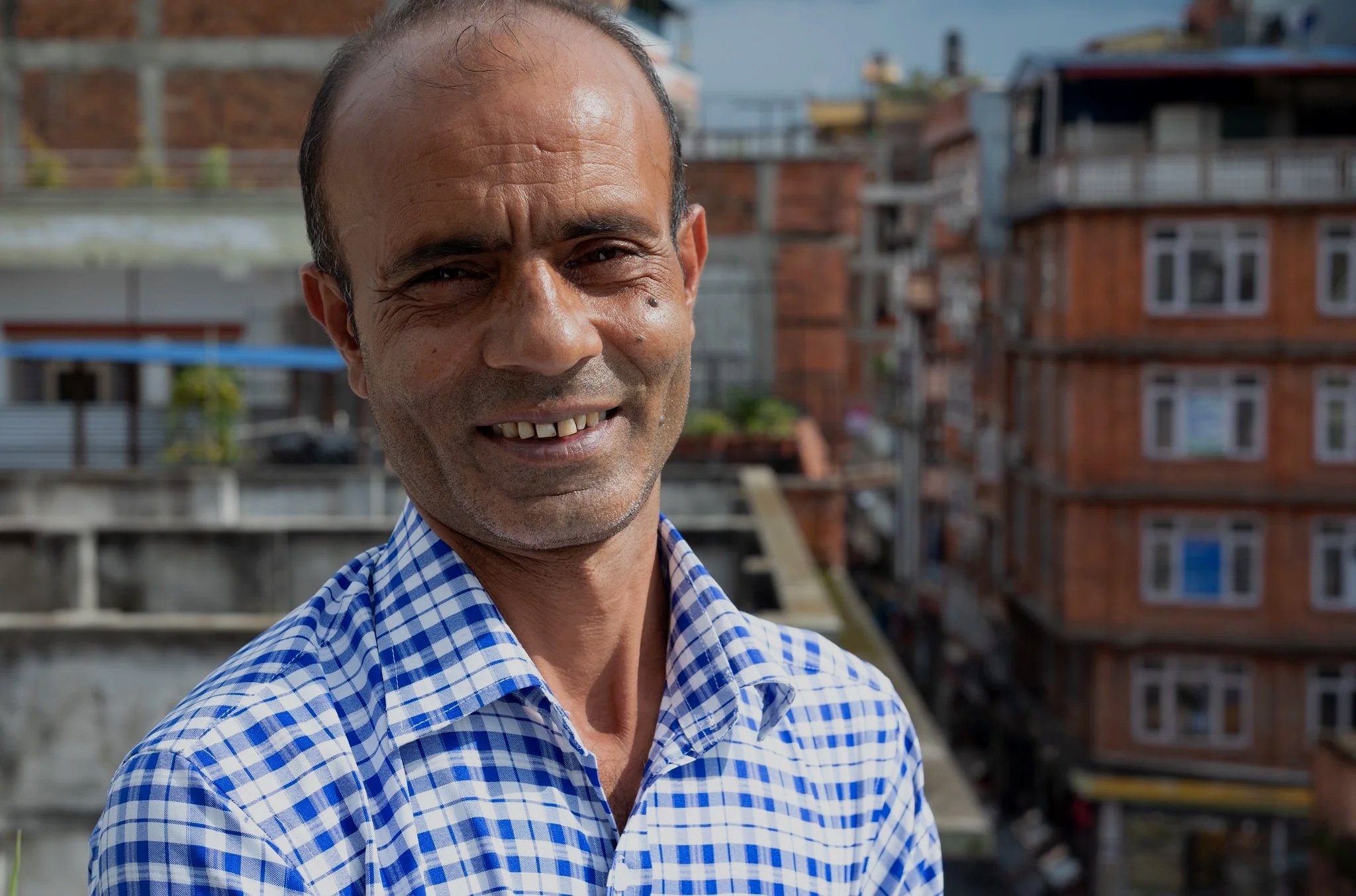 Smiling man in a checkered shirt standing outdoors with apartment buildings in the background.