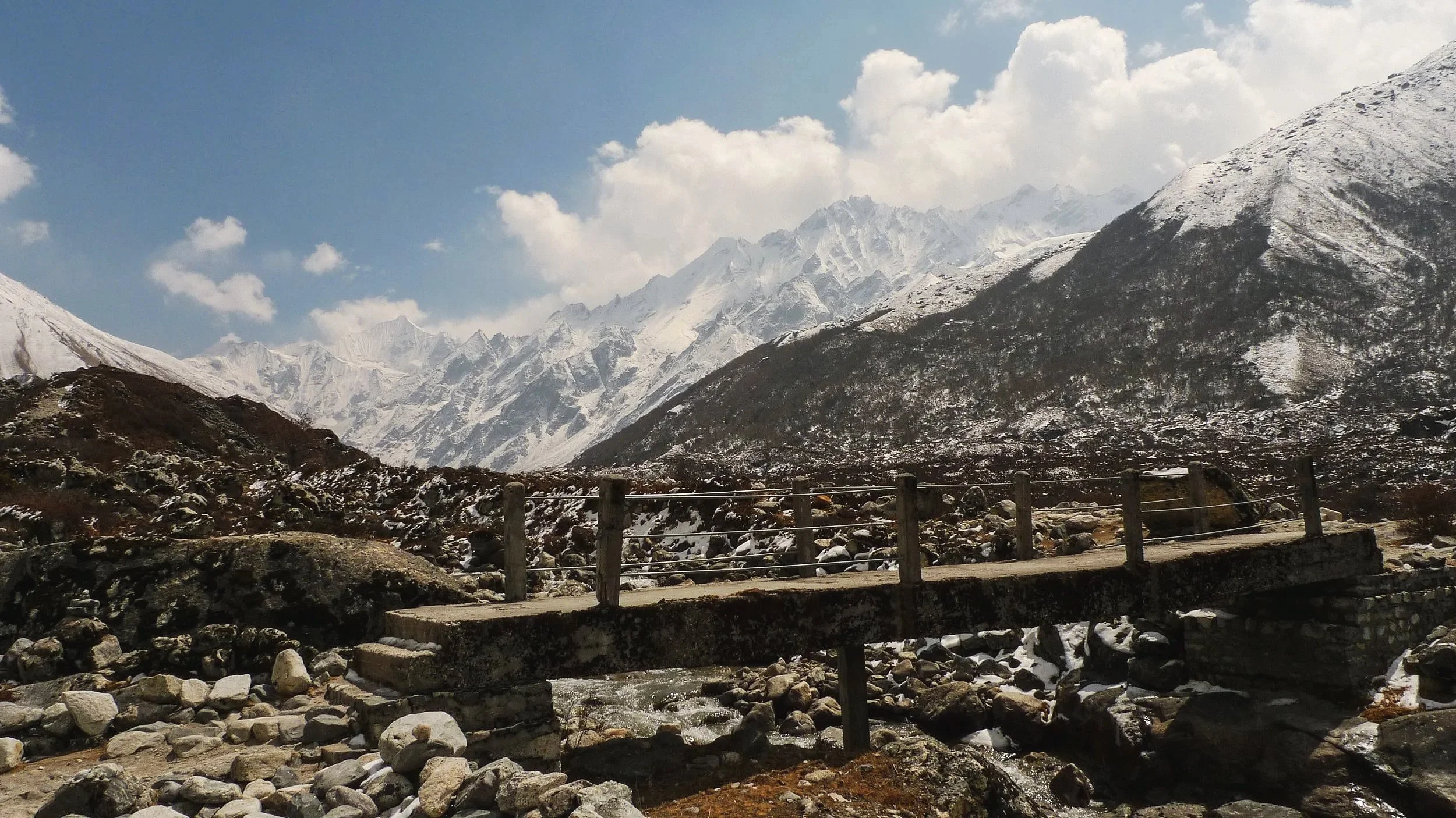 A mountain landscape with snow-capped peaks, a wooden bridge crossing a rocky stream, and a partly cloudy sky, in Langtang National Park.