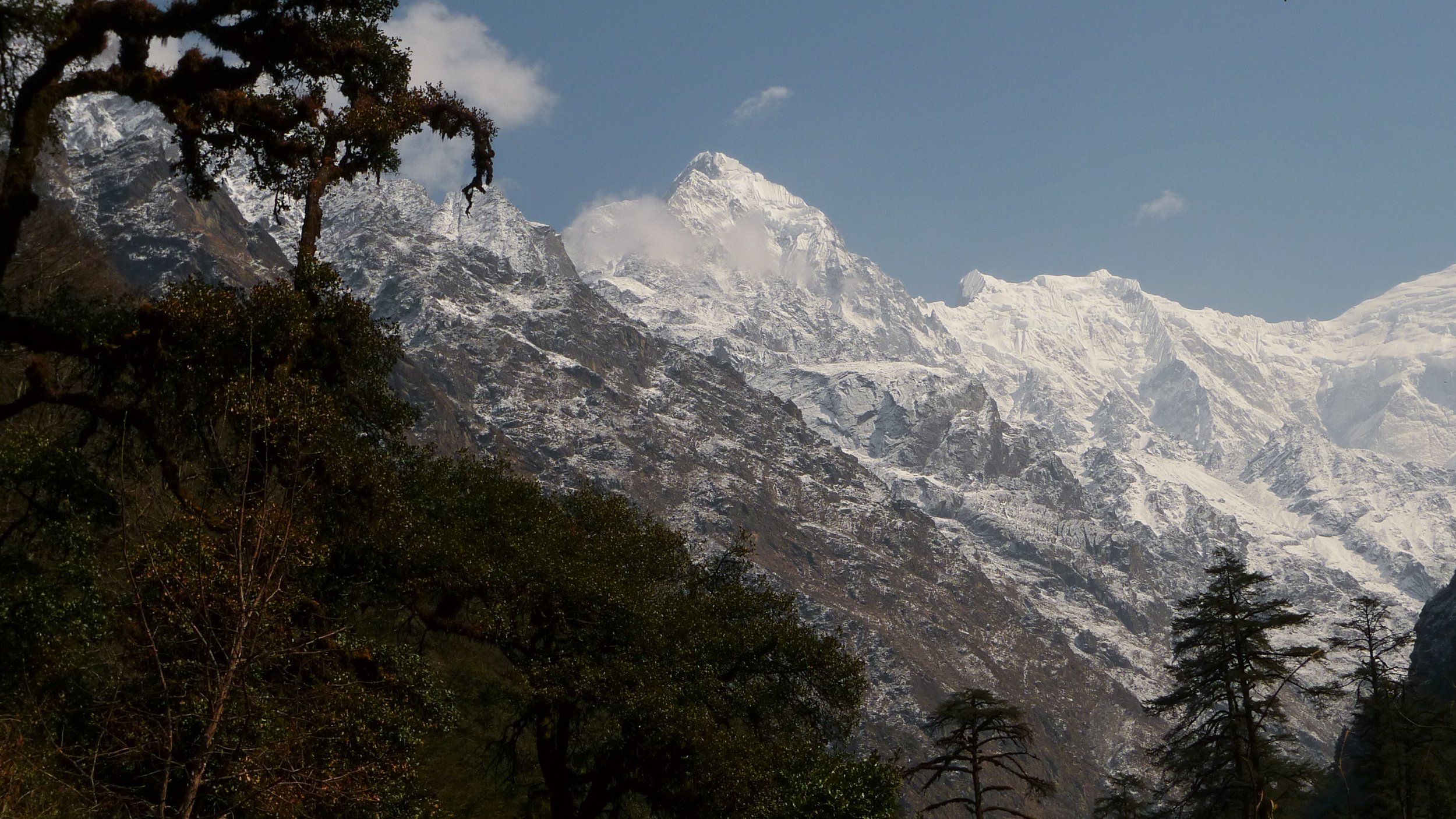 Snow-capped mountain peaks with trees in the foreground and a partly cloudy sky.