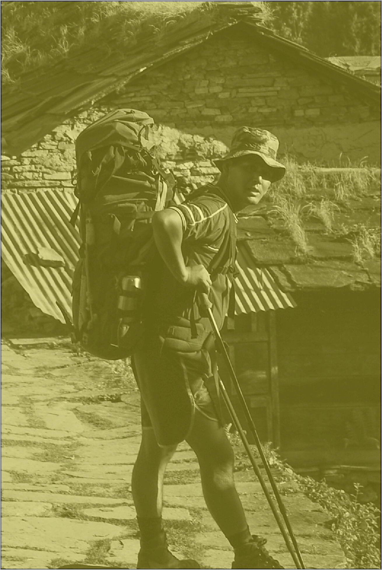 A man with a backpack and hiking poles standing on a rocky path in front of a rustic building with a stone chimney and metal roof.