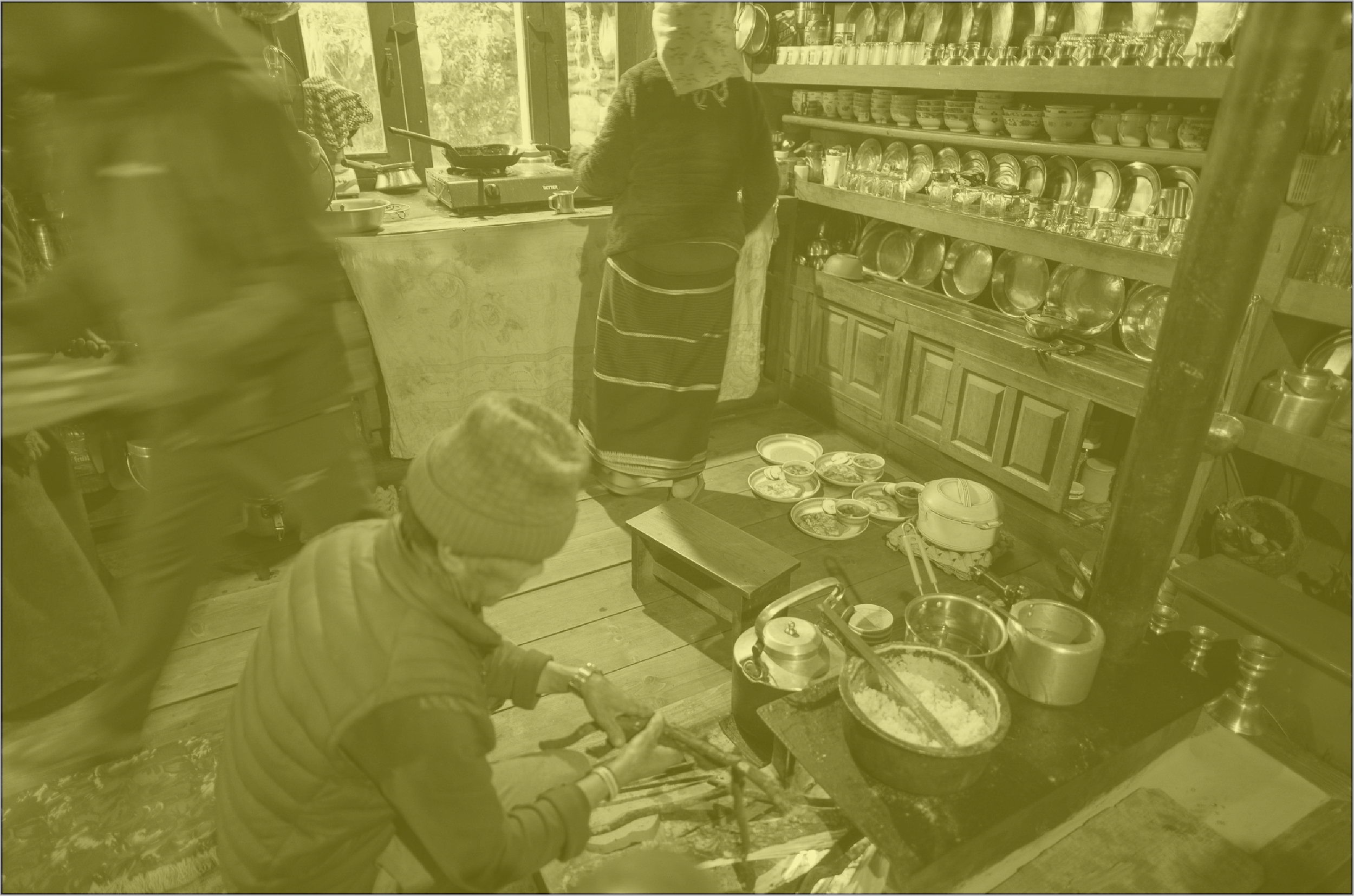 A person wearing a striped jacket and a beanie preparing food on a wood surface with cooking utensils, bowls with food, and a black stove nearby in a kitchen, while a woman in traditional clothing and an apron stands at the counter preparing food and shelves filled with bowls and utensils are in the background.