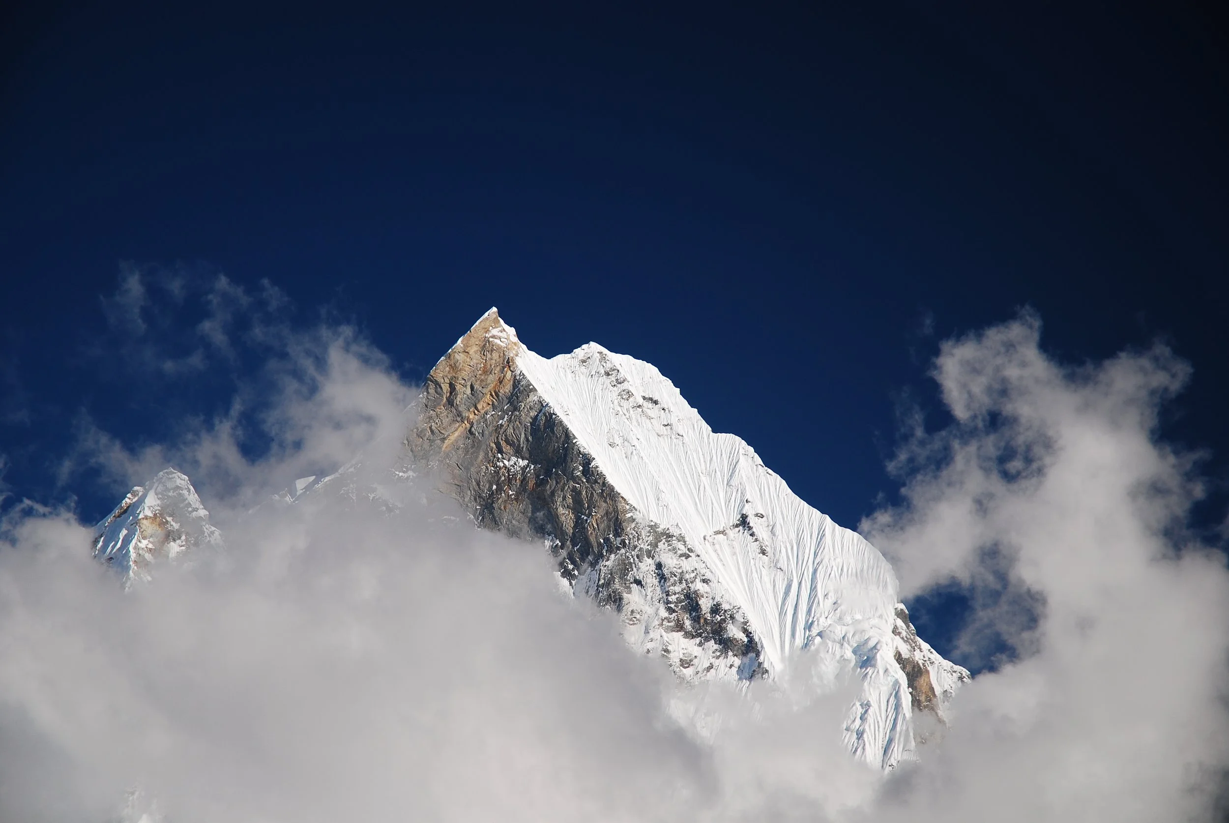 Snow-covered mountain peak rising above clouds against dark blue sky.