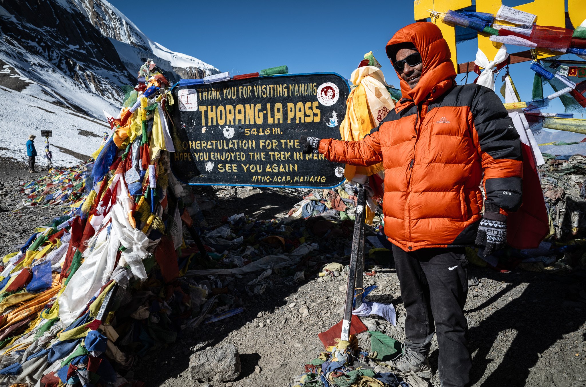 A man dressed in an orange jacket and black pants standing next to a black sign with yellow and white text at Thorung-La Pass, 5416 meters high, thanking visitors and congratulating trekkers in Manang, Nepal. The sign is decorated with colorful prayer flags and is situated in a snowy mountainous landscape.